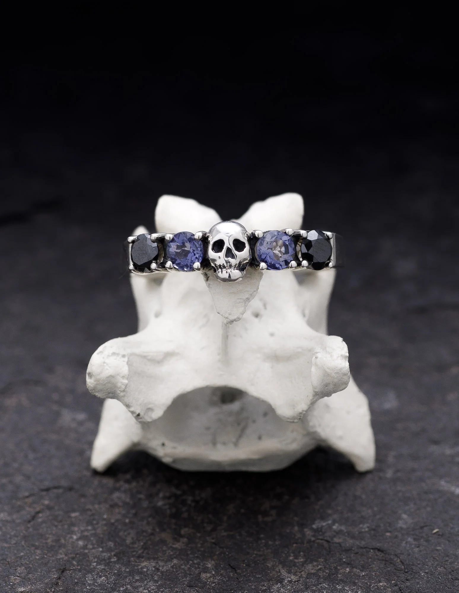 Ring with a skull in the center, flanked by two purple sapphires and two black stones, displayed on a white anatomical model of a human vertebra.