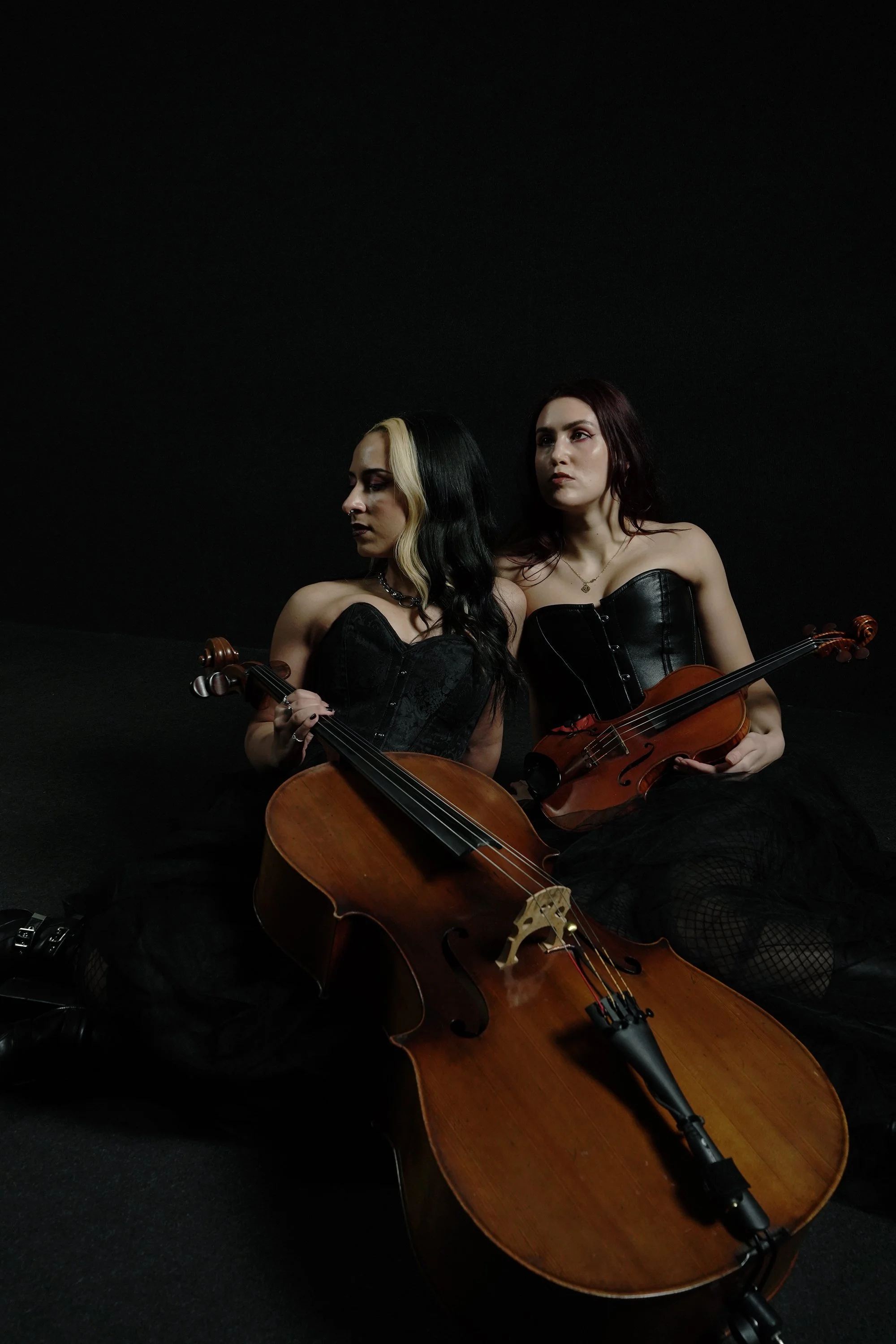 Two women dressed in black corsets sitting on the floor against a black background, each holding a violin, with a large cello lying in front of them.
