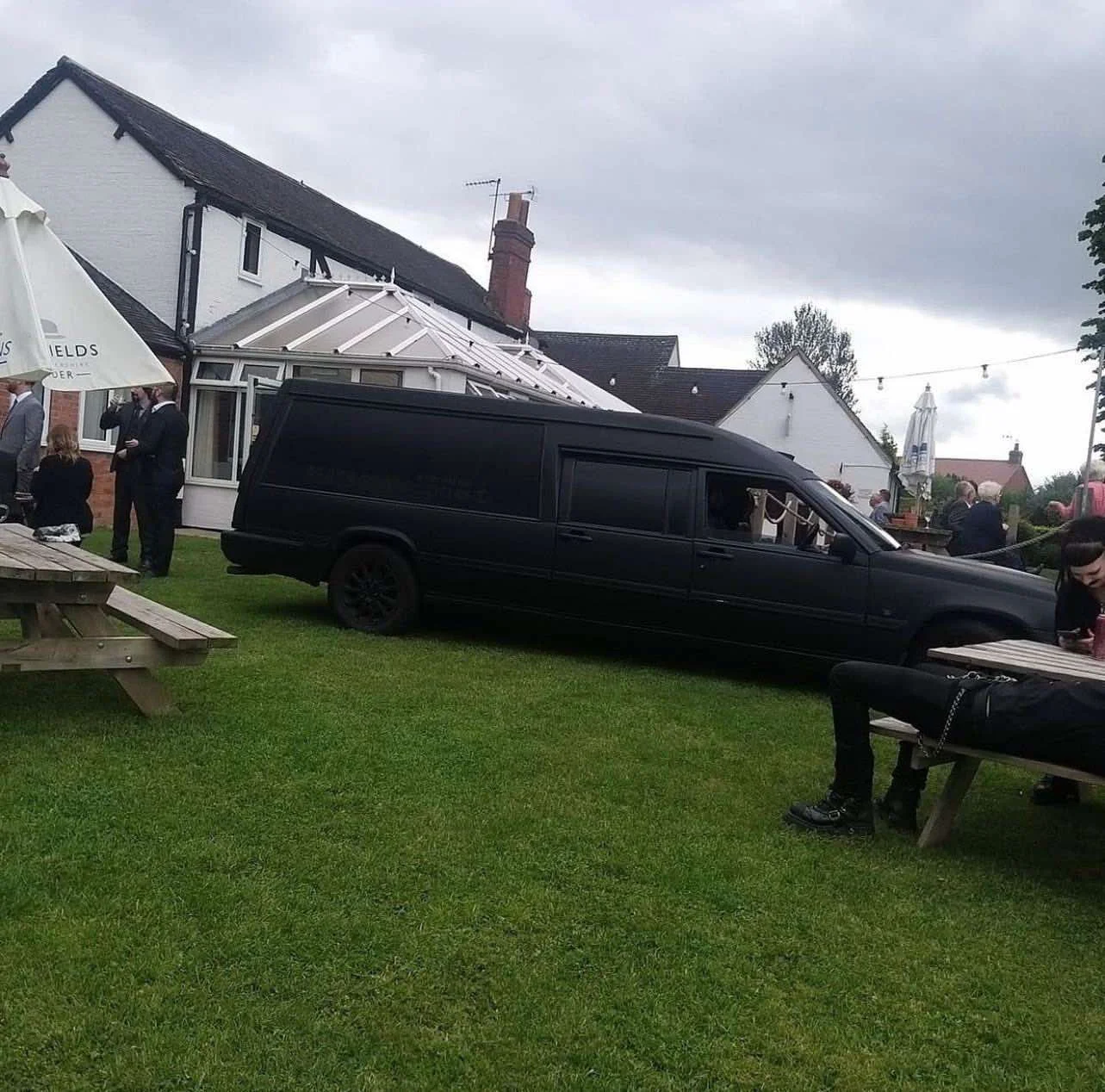 A black hearse parked on a grass lawn during an outdoor gathering with people dressed in formal attire near houses under a cloudy sky.