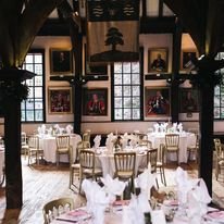Elegant dining room with round tables set with white tablecloths, napkins, and glassware, and gold chairs, featuring large windows and decorative shelves