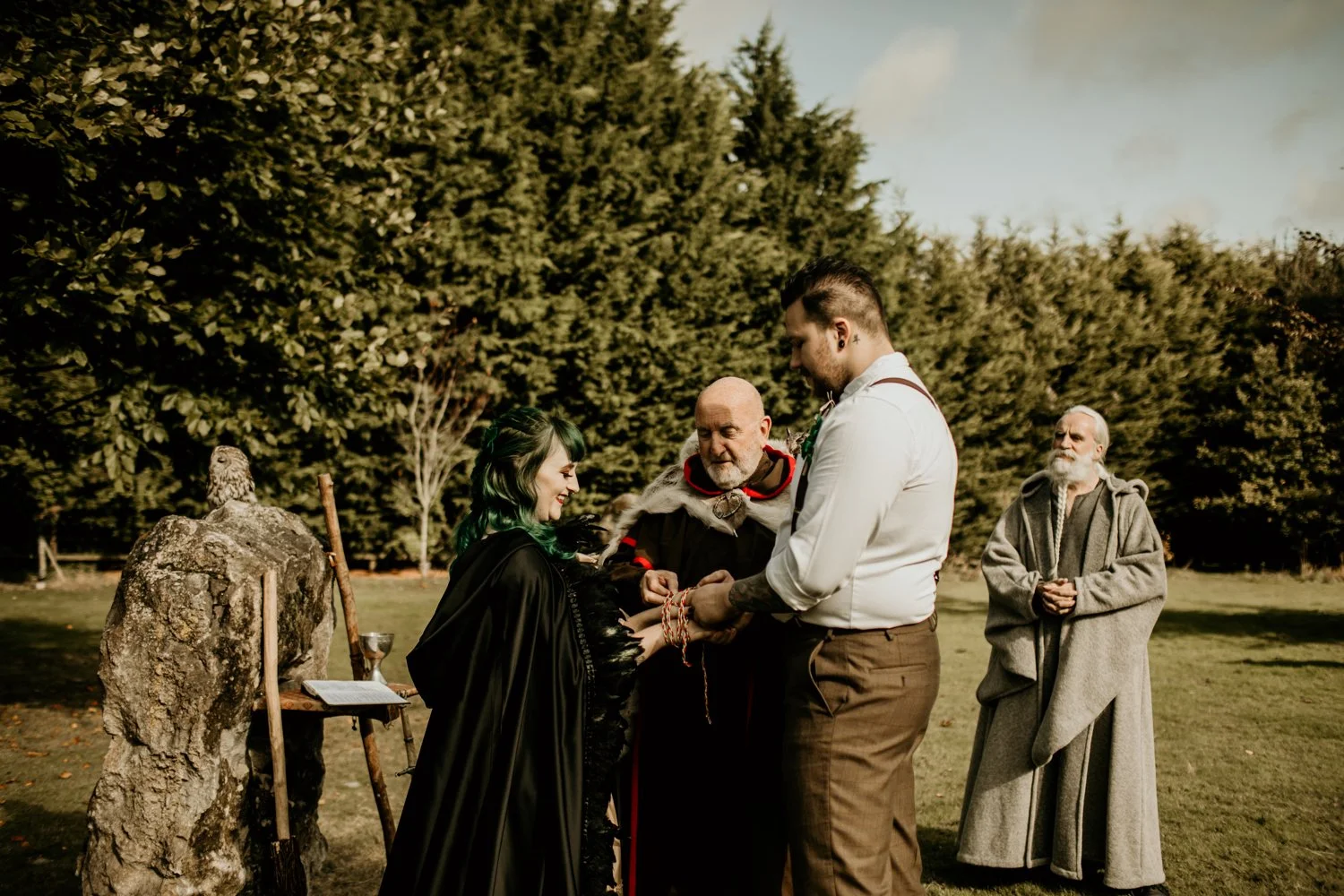 A wedding ceremony taking place outdoors with four people: a bride with green hair, a groom, a priest or officiant, and a woman dressed in a hooded robe resembling a monk. The couple is holding hands and looking at each other, while the woman in the 