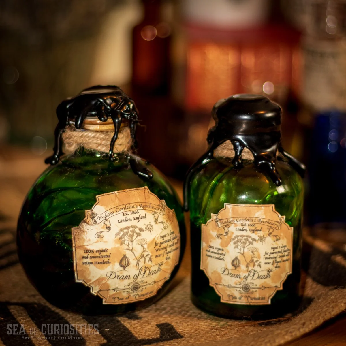 Two green bottles labeled 'Dram of Death' with black wax seals on the caps, placed on a surface with a blurred background of warm-colored objects.