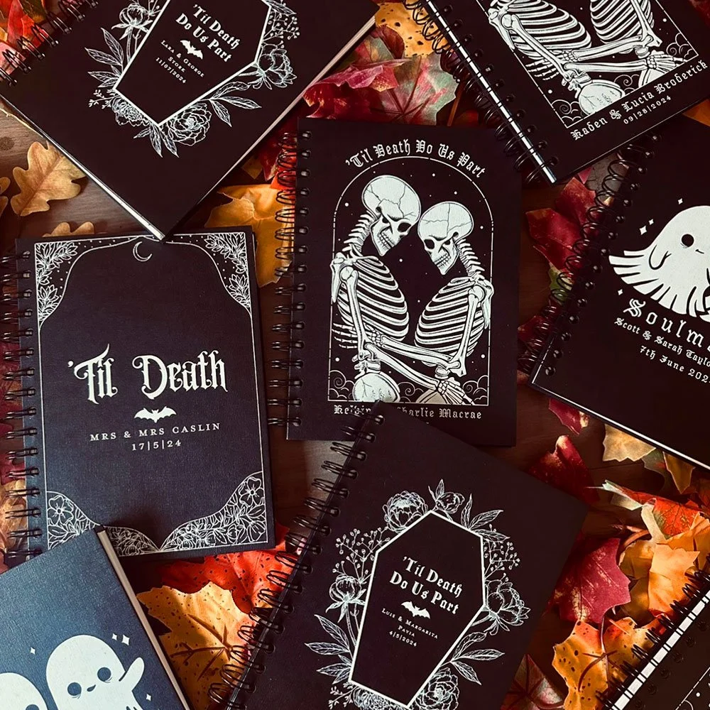 Multiple black notebooks with white skeleton and Halloween-themed designs scattered on autumn leaves.