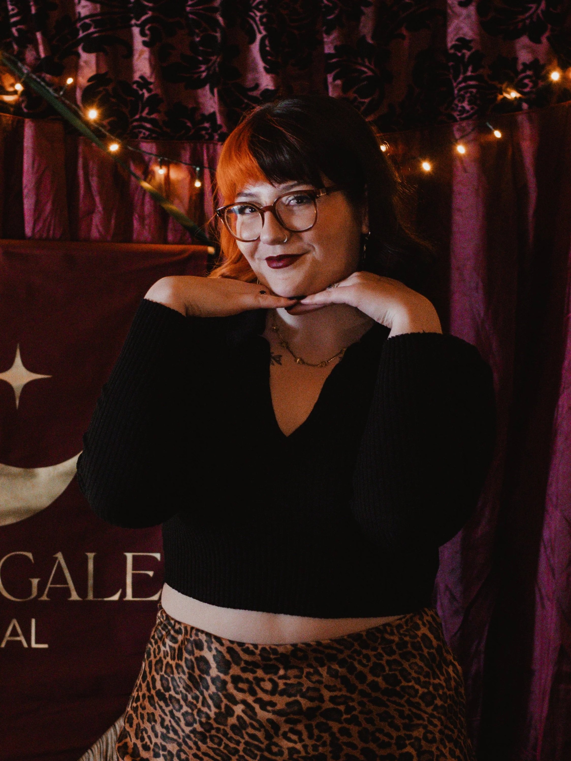 A young woman with red hair, glasses, and dark lipstick posing with her hands under her chin, smiling in front of a dark purple curtain with string lights and a partial banner.