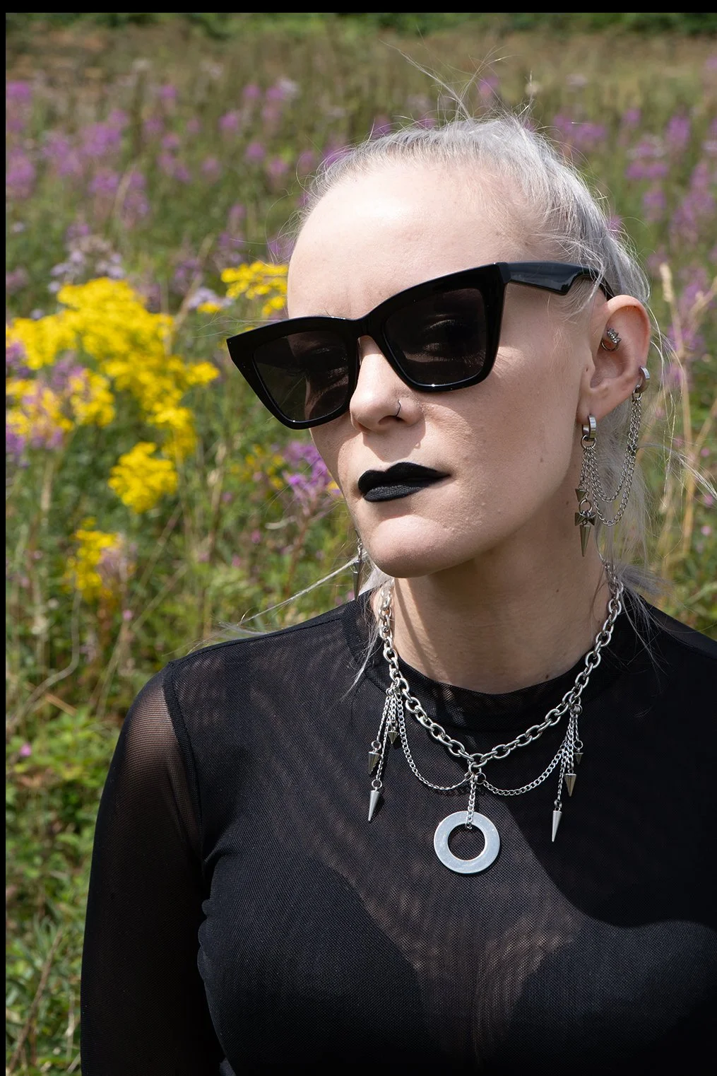 A woman with platinum blonde hair wearing large black sunglasses, multiple silver chains with pendants, and black lipstick, standing outdoors in front of a colorful wildflower field.