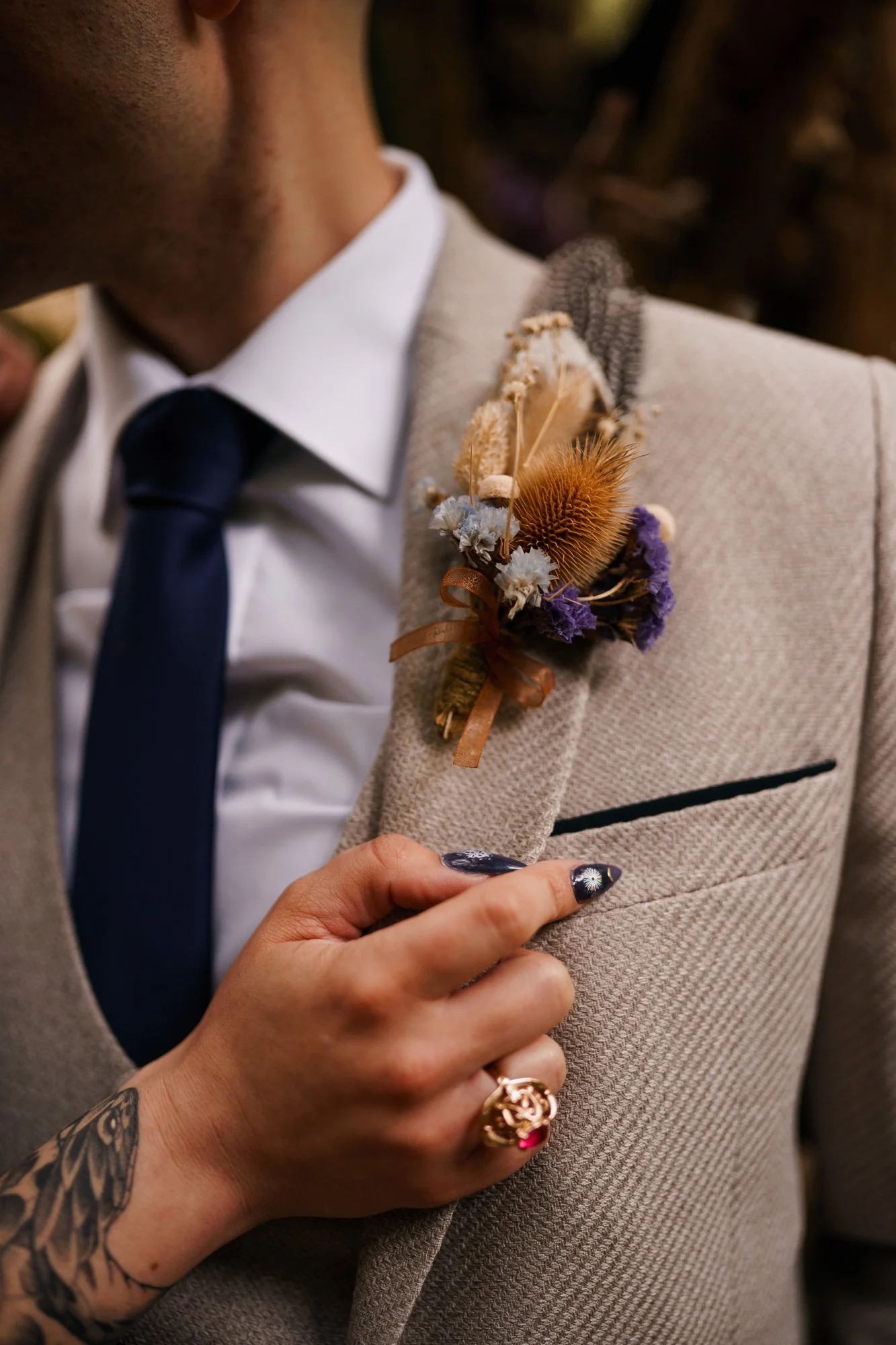 Close-up of a man in a beige suit and dark tie with a floral boutonniere on his lapel, and a woman's hand adjusting his jacket. The woman's hand has decorated nails and a ring.