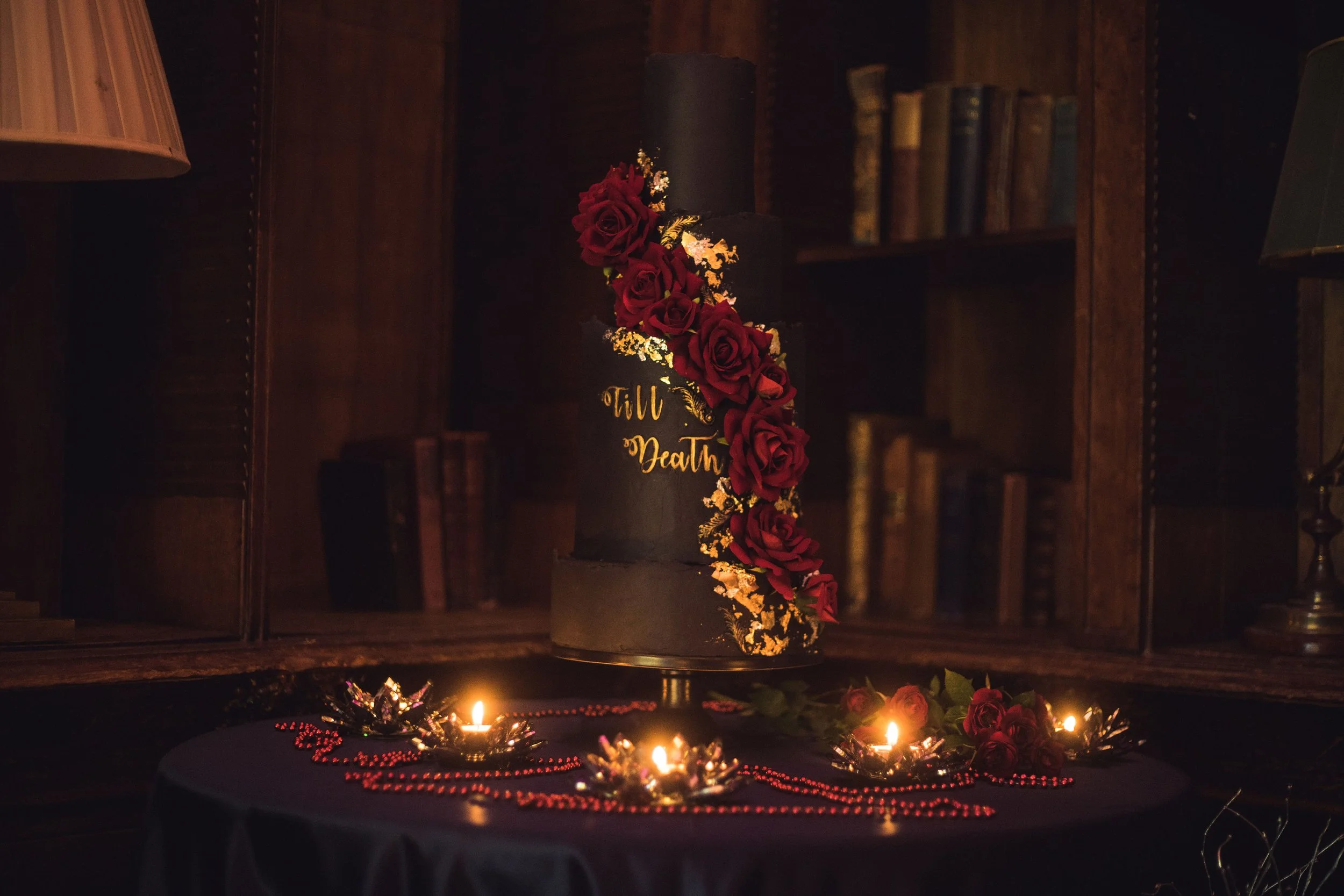A tall black cake decorated with red roses and gold accents, placed on a dark table with candles and red beads, in a wooden room.