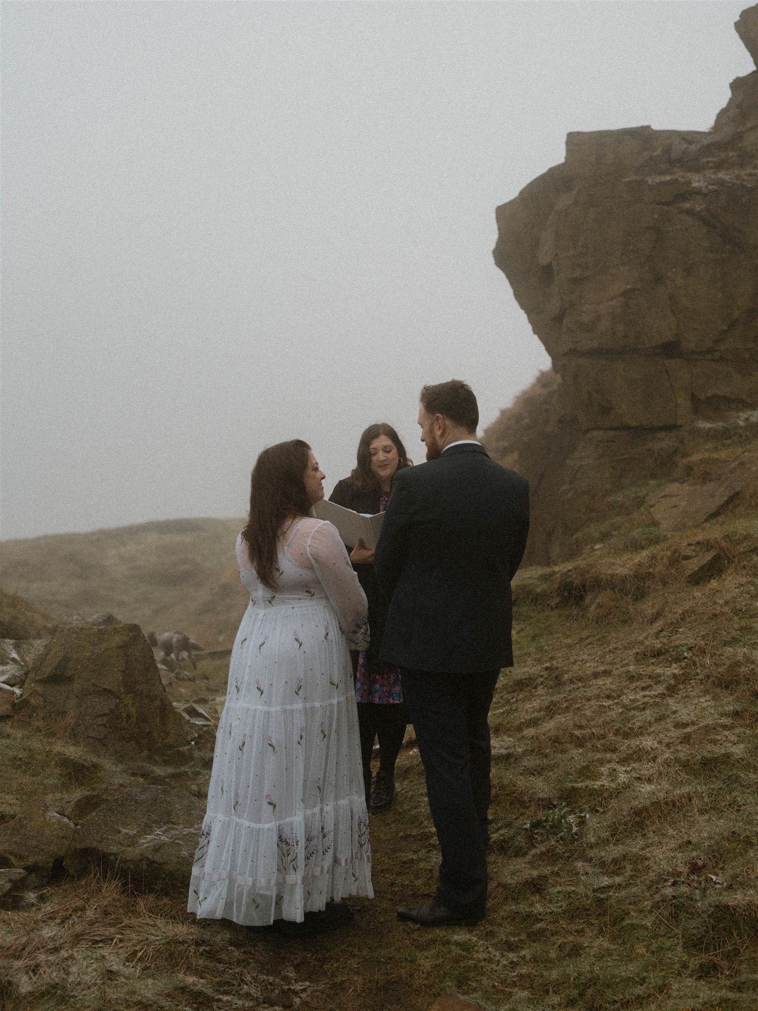 A couple getting married outdoors on a foggy day, standing on a dirt path between rocks and grass, with a woman officiant reading from a book.