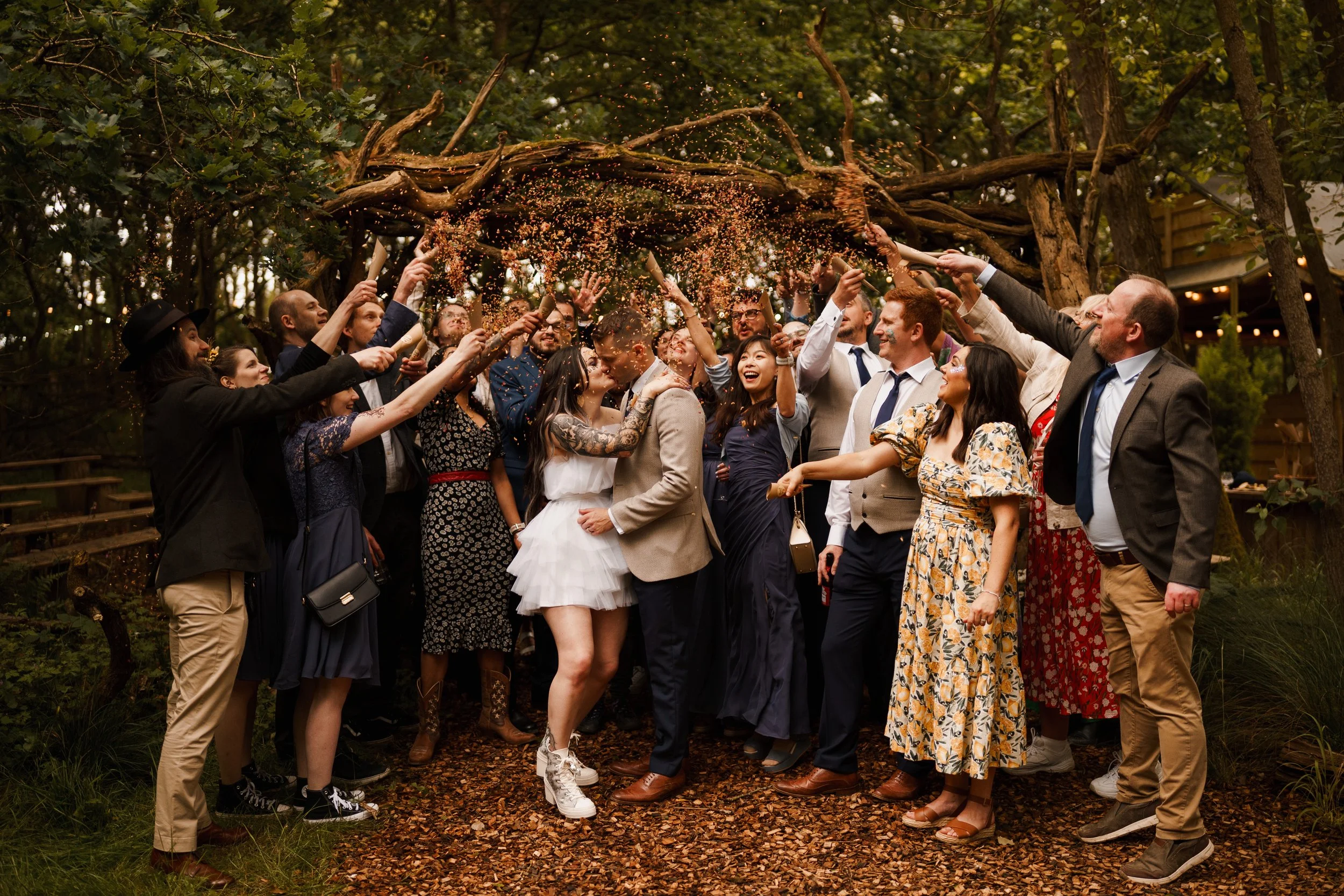 A group of people celebrating a wedding outdoors, throwing confetti over a couple who are kissing in the center.