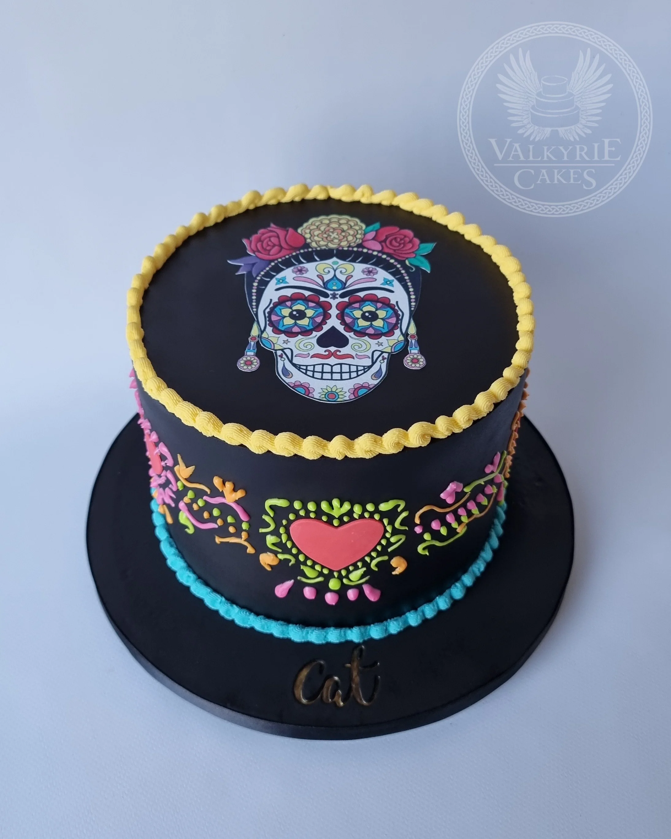 Two-tier cake decorated with colorful sugar skull, flowers, hearts, and floral patterns, with 'cat' written on the cake board.
