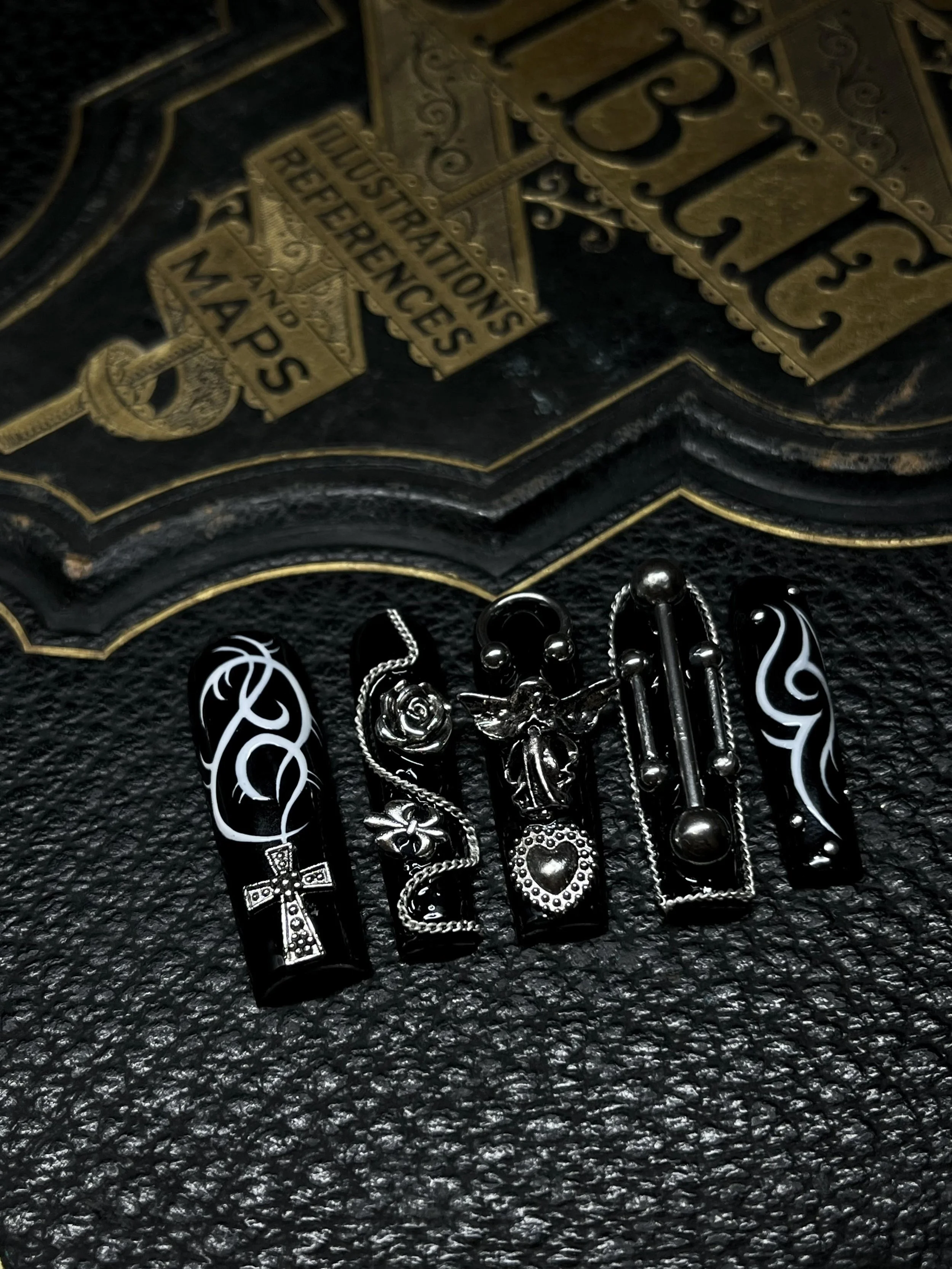 Five black and silver decorative nail polish bottles with various charms, set on a textured black surface with a vintage-style sign reading 'Blues' and a yellow traffic signal sign in the background.