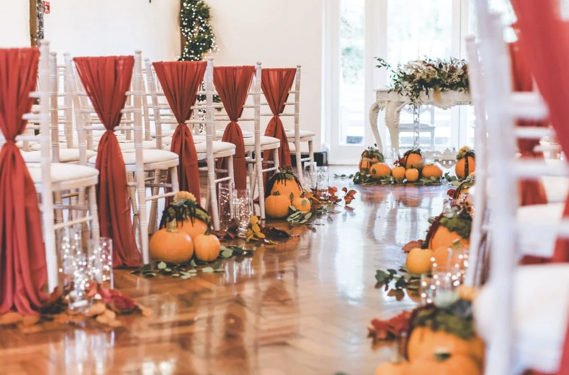 Decorated indoor space with white chairs draped with red fabric, pumpkins, fall leaves, and string lights for a seasonal event or celebration.