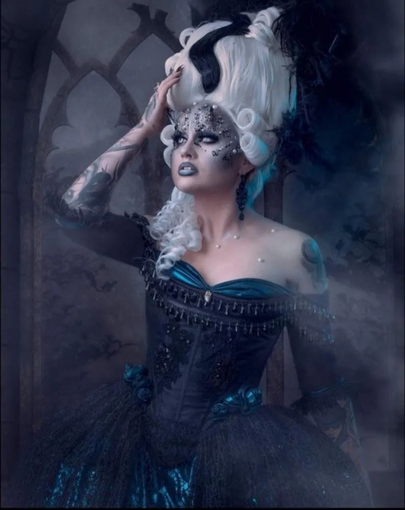 A woman with elaborate white and black hair styled in curls wearing a dark, ornate dress with blue highlights, surrounded by mist or fog, with gothic-style window in the background.