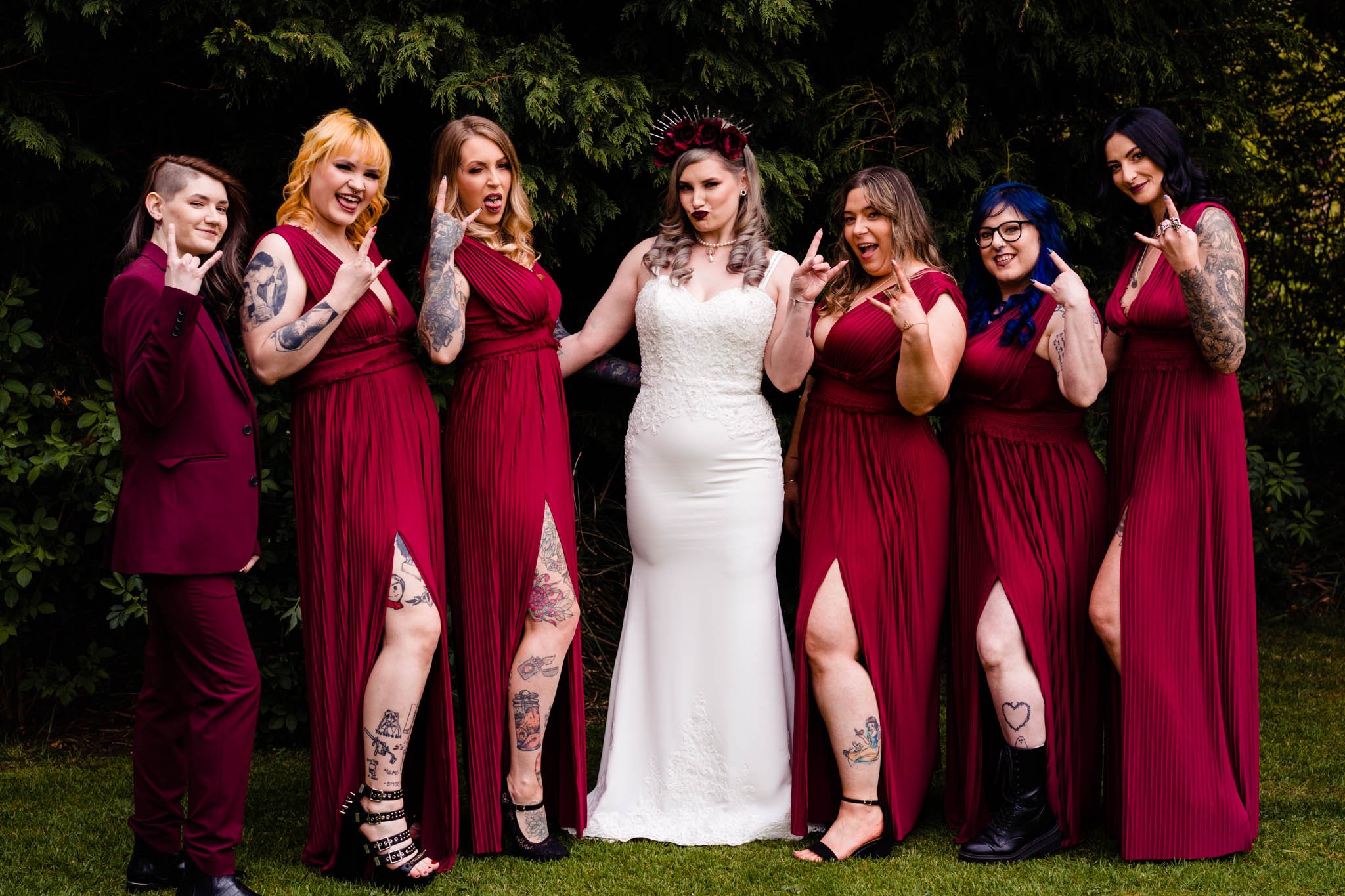 Group of women, including one bride in a white gown and six bridesmaids in matching burgundy dresses, standing outdoors with greenery in the background.
