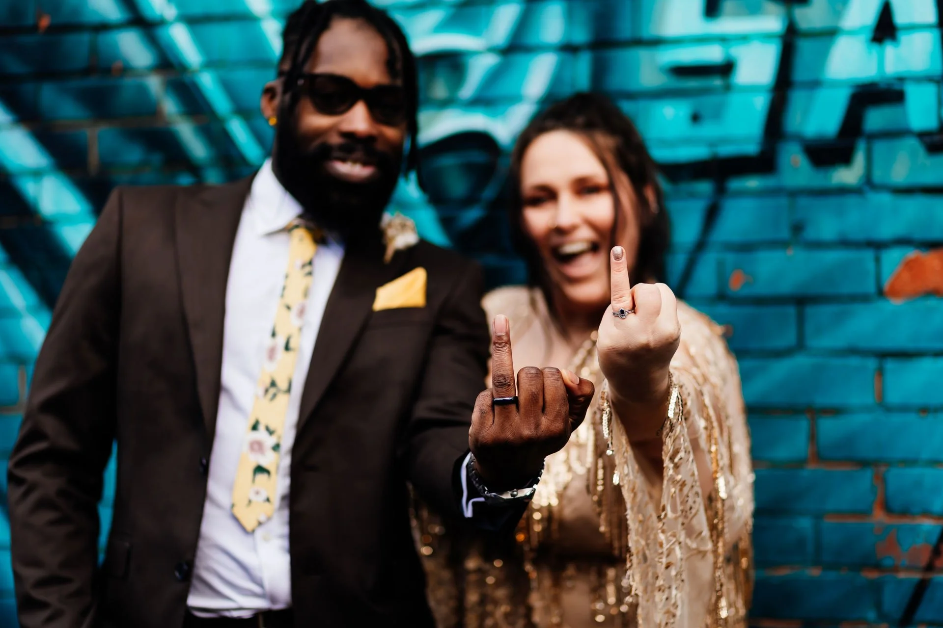 Two people standing in front of a blue graffiti wall, holding up their middle fingers to the camera, smiling.