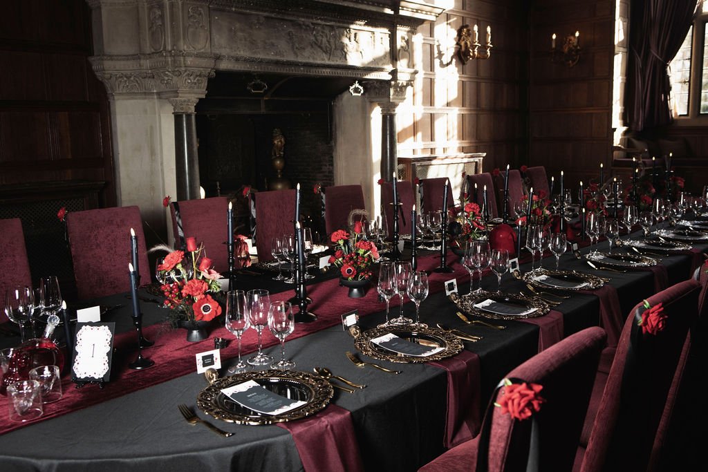 A long dinner table set for a formal event in a dark wood-paneled room with a stone fireplace. The table is covered with black and burgundy tablecloths, and decorated with black candlesticks, red flowers, and elegant place settings including gold-rim