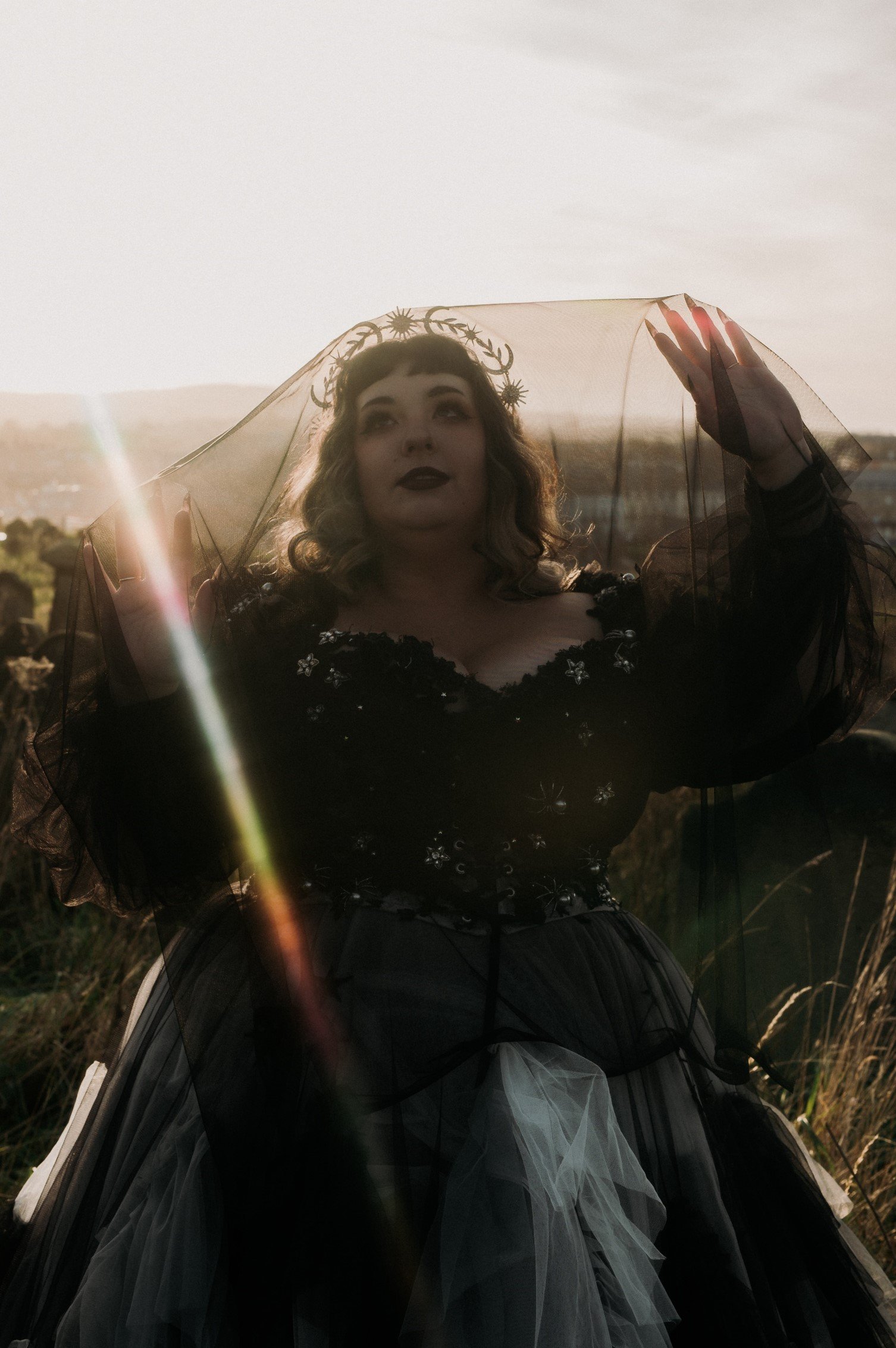 A woman dressed in a black and white Gothic-style gown with floral embroidery, standing outdoors at sunset with her arms raised, wearing a veil and a headpiece with cosmic motifs.