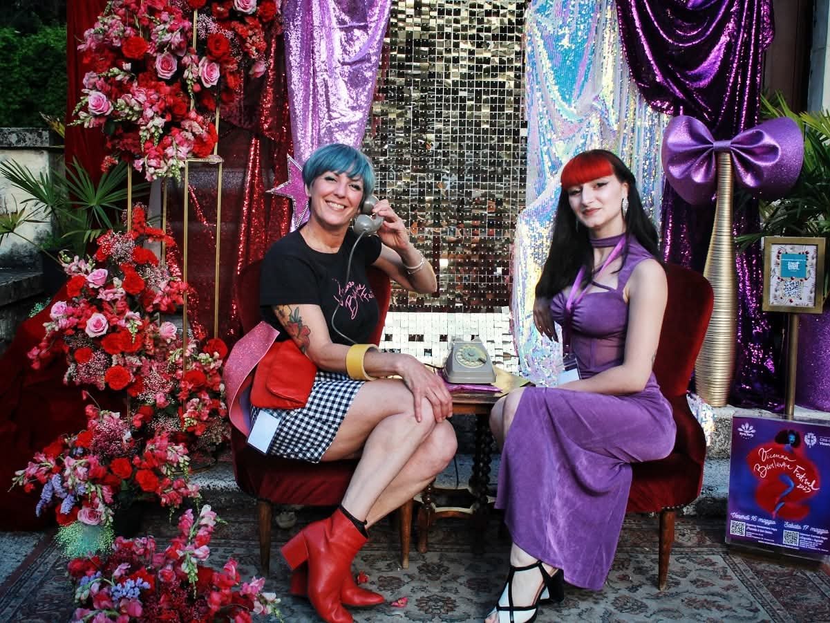 Two women sitting on red velvet chairs in front of a colorful backdrop with flowers, sequins, and ribbons. One woman with blue hair is holding a vintage rotary phone, smiling, dressed in a black t-shirt, checkered skirt, and red boots. The other woma