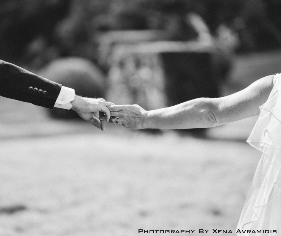 A black and white photo of a person in a wedding dress holding hands with someone in a suit, with a blurred outdoor background.