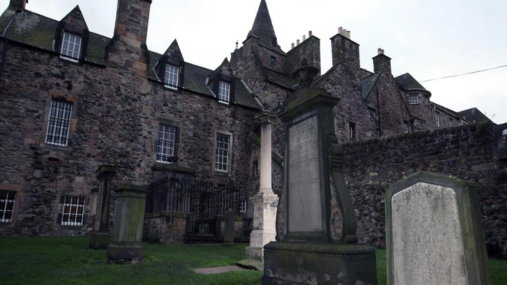 Old graveyard with tombstones and a historic stone building with chimneys and steeples.