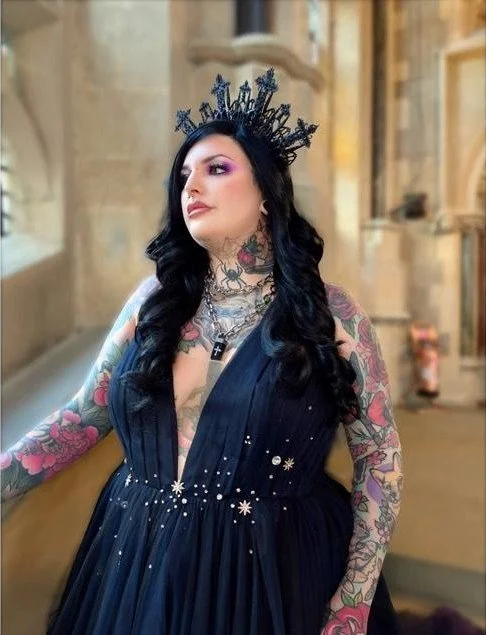 Woman with long black hair and colorful tattoos wearing a dark blue dress and a decorative headpiece, standing indoors.