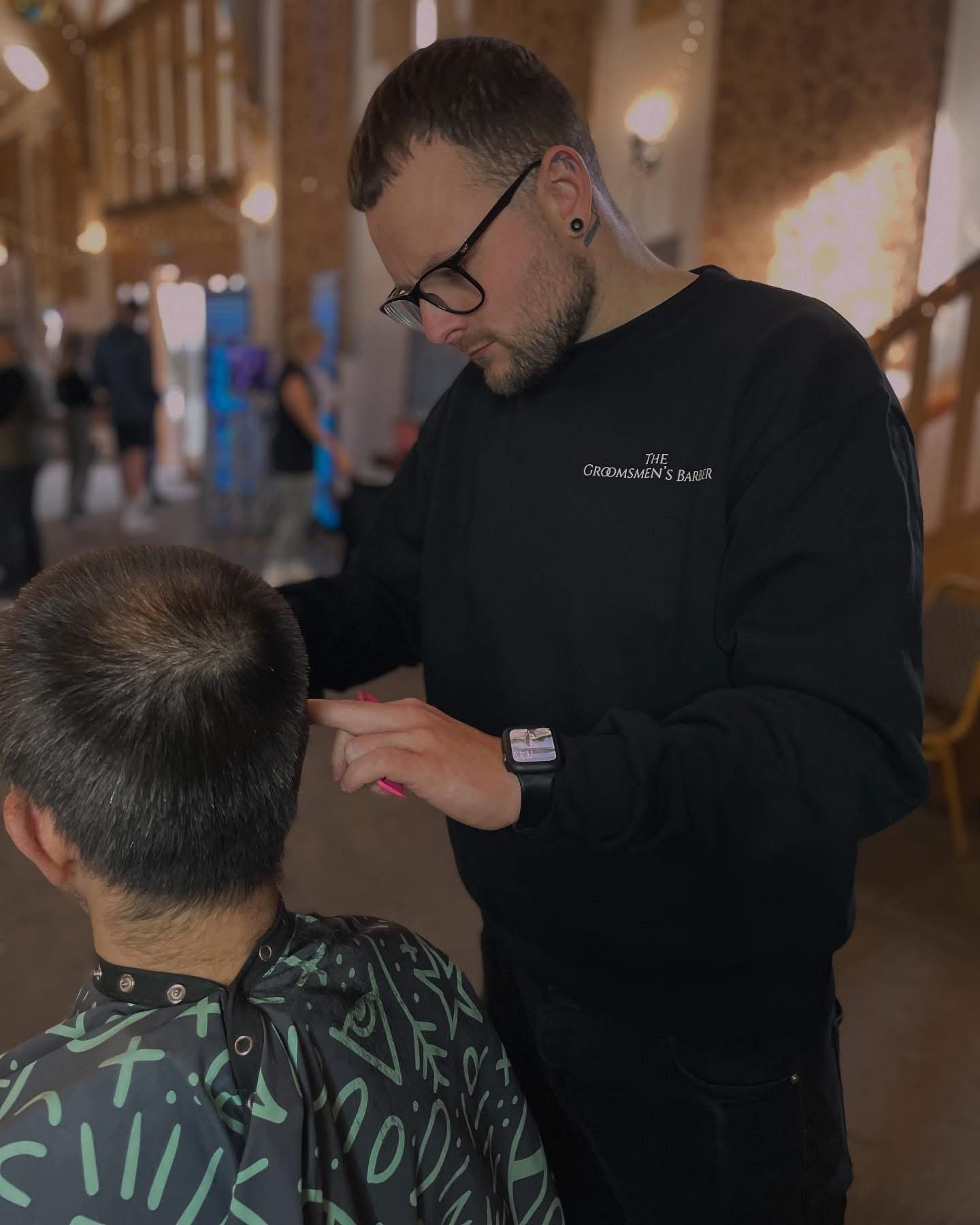 A barber with tattoos, glasses, and a beard cuts a customer's hair inside a barbershop. The barber wears a black shirt that reads 'The Groomsmen's Barber' and an Apple Watch. The customer has short, dark hair and is wearing a cape with geometric and 