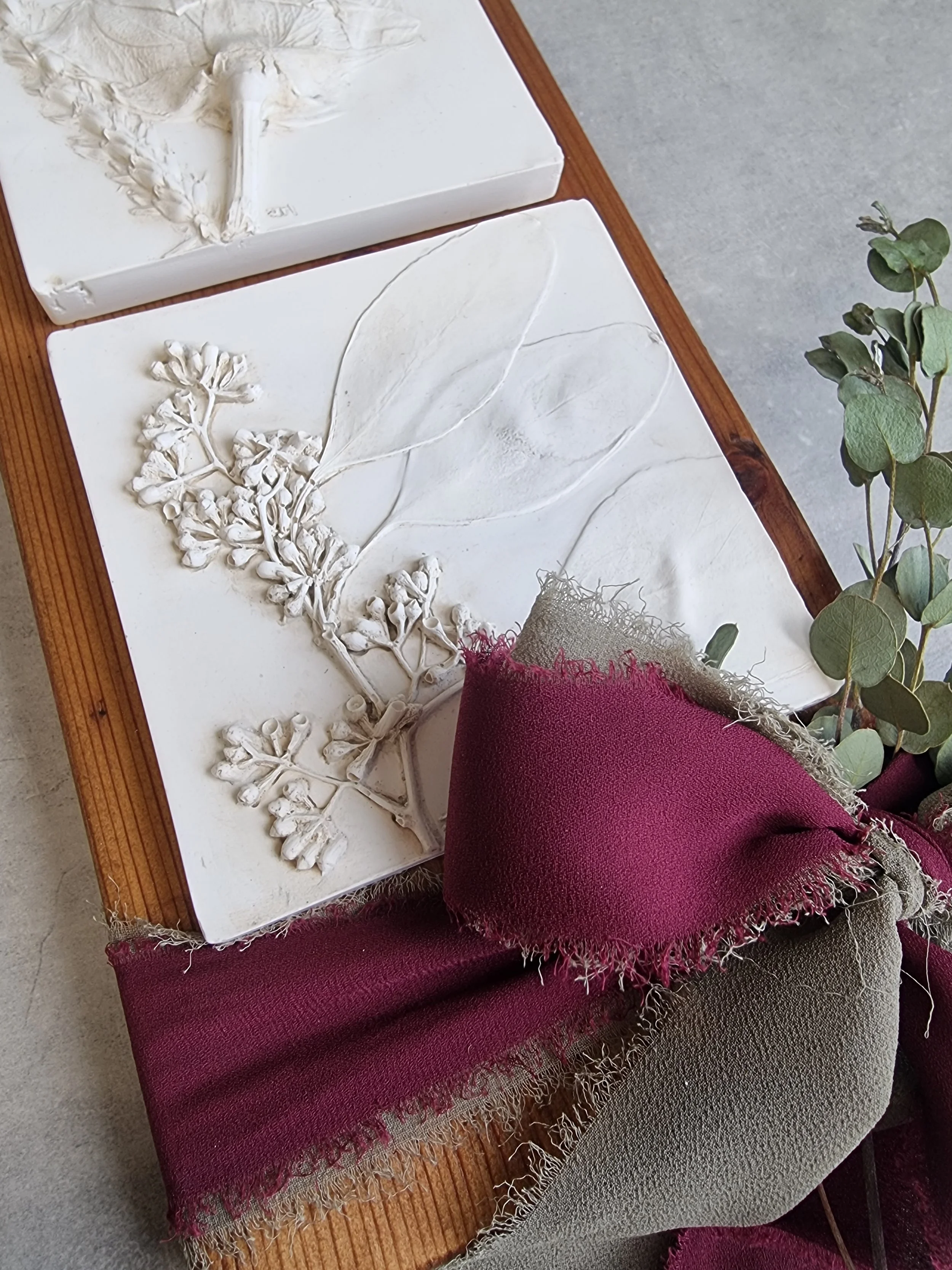 Two white ceramic plaques with raised botanical designs, one featuring a branch with small flowers and leaves, are placed on a wooden table. A branch with green eucalyptus leaves and a piece of burgundy fabric with frayed edges are also on the table.