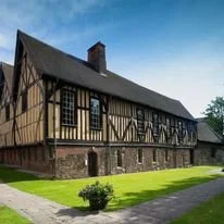 Historical Tudor-style building with black wooden beams and white walls, set on a grassy lawn under a clear blue sky.