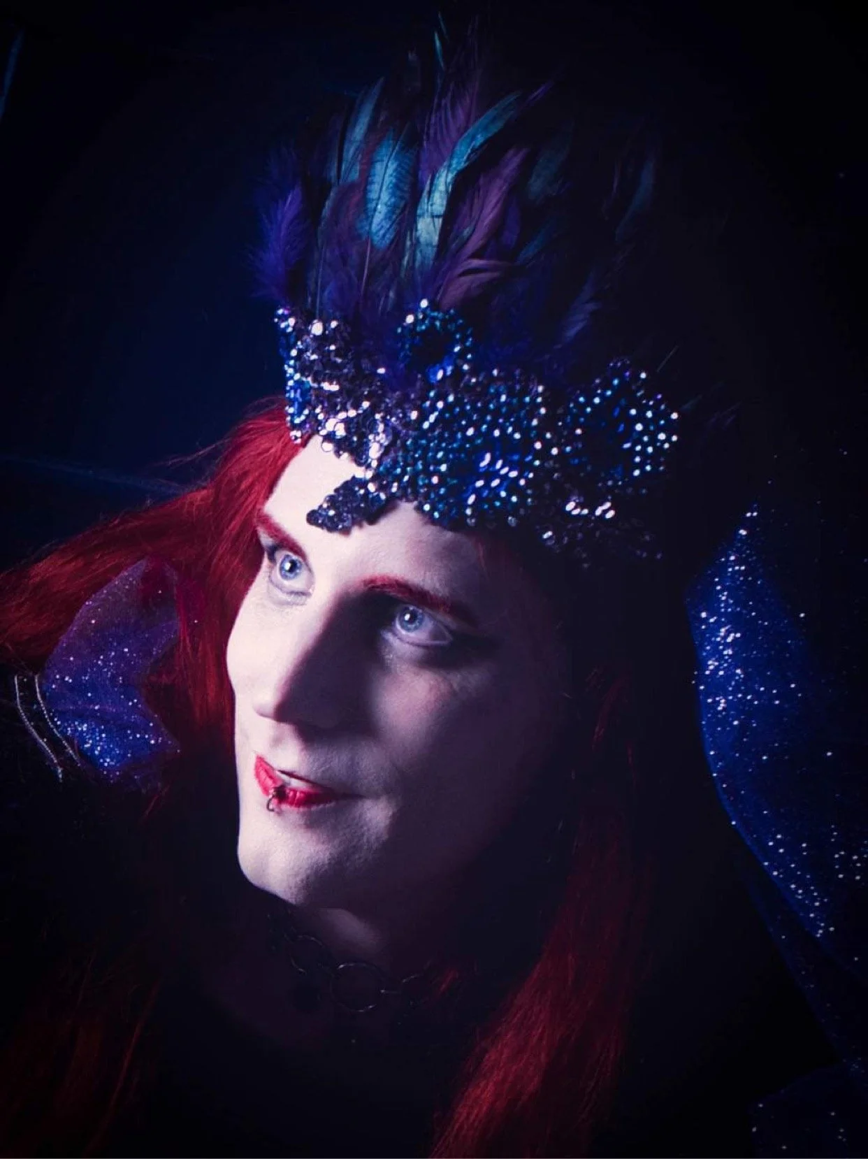 A person with red hair and pale skin wearing a decorated costume with blue feathers and sparkles, looking upward.