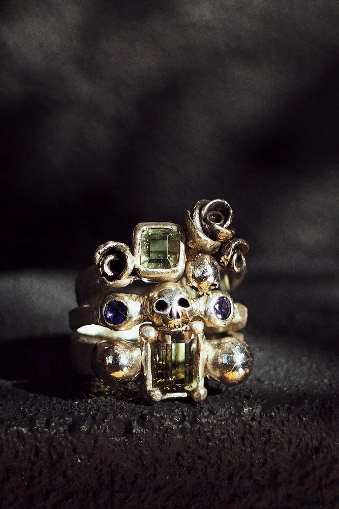 Silver ring with skull design, featuring green and purple gemstones, against a black background.