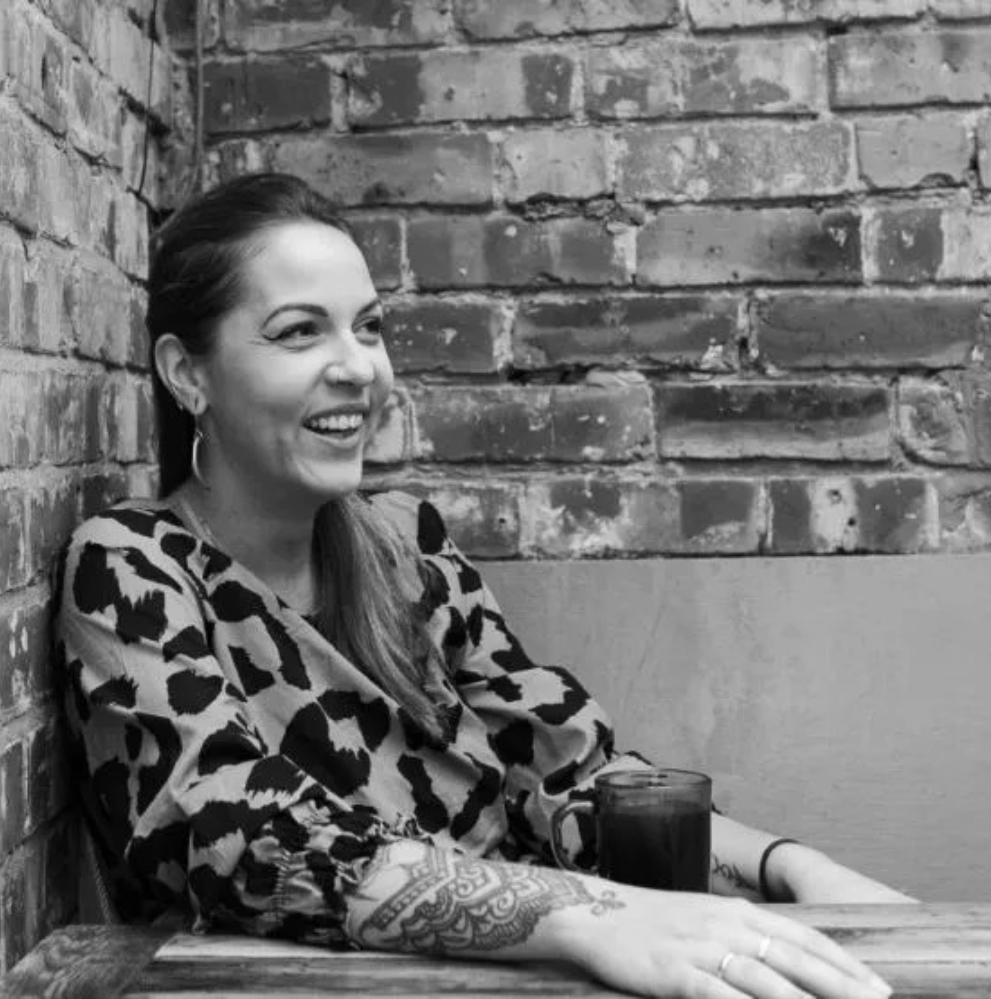 A woman with a tattoo on her arm smiling and sitting at a table with a mug, against a brick wall.