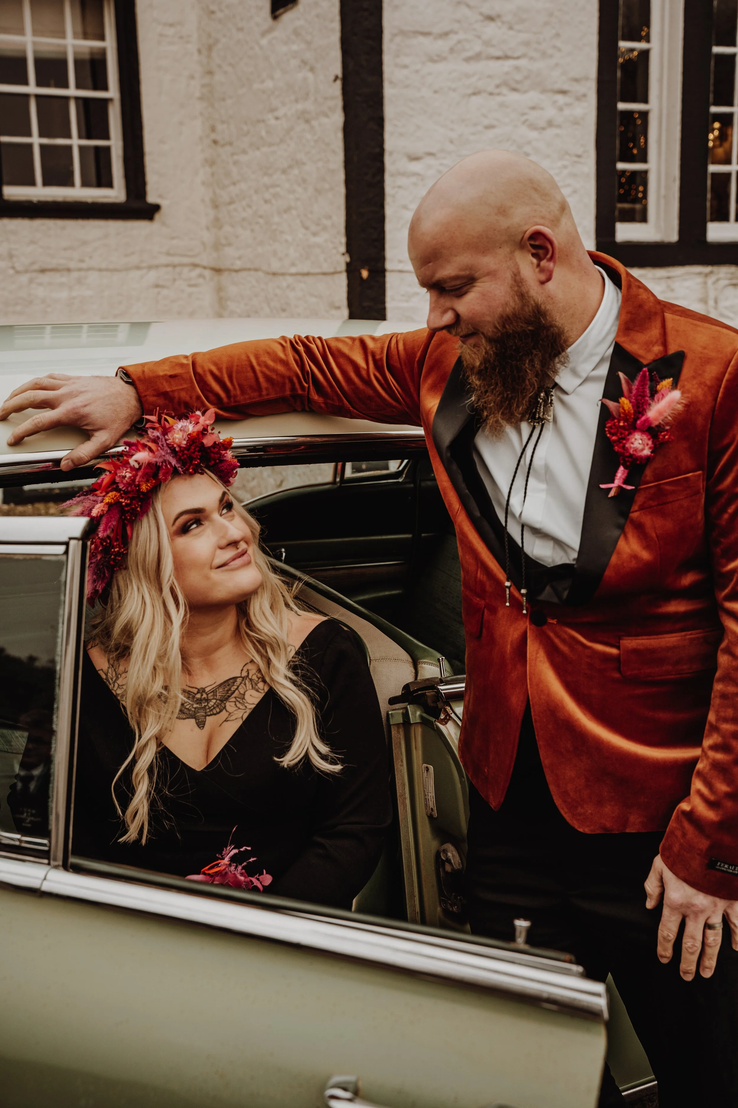 A woman with long blonde hair and a tattoo on her chest sitting in a vintage car, looking up at a man with a beard and a shaved head leaning on the car, both dressed in formal attire. The woman is wearing a black dress and a pink floral crown, while the man is wearing an orange velvet tuxedo jacket with a pink floral boutonniere.