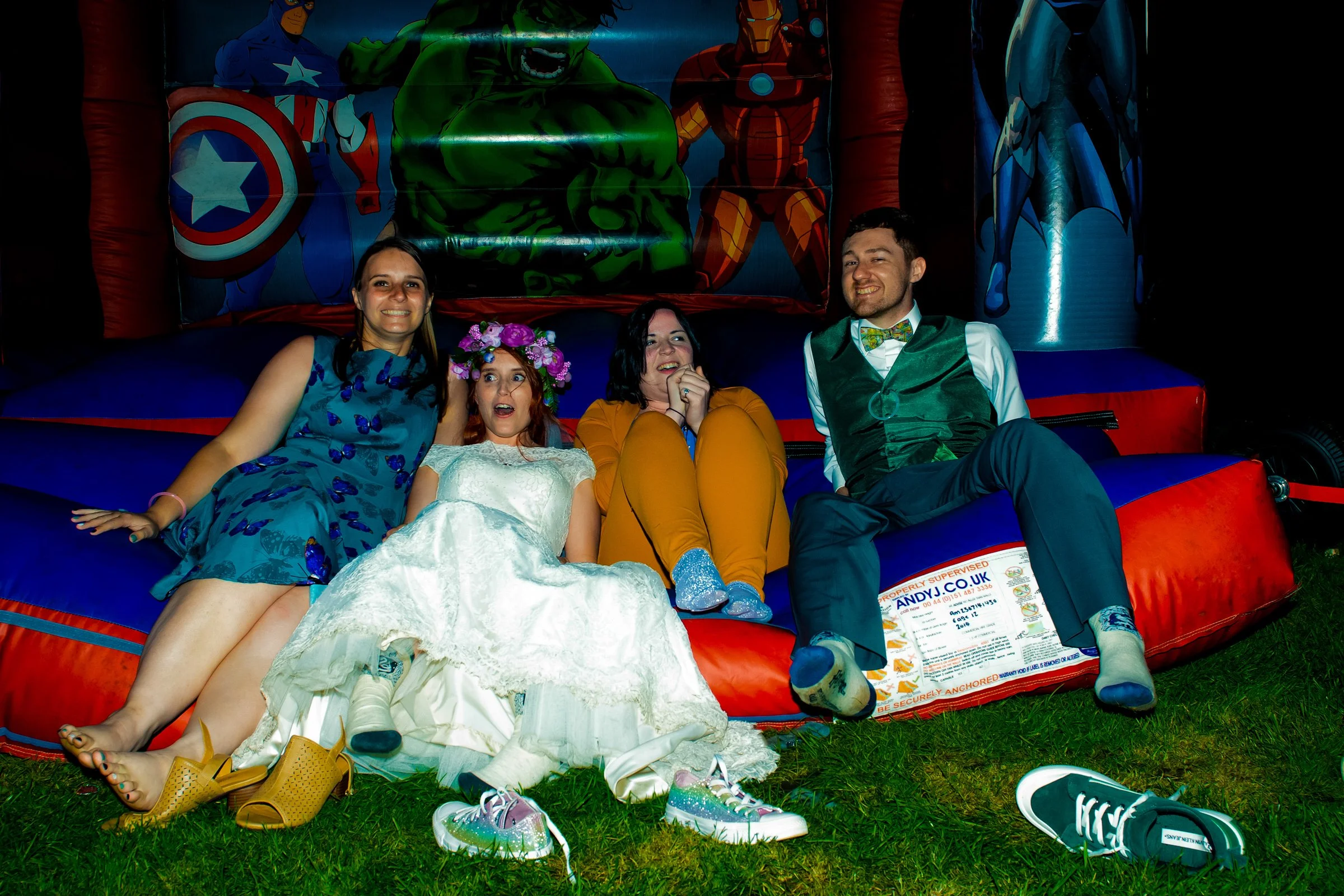 Group of five people sitting on a bouncy castle at night. They are smiling and dressed in colorful clothes, with a superhero-themed backdrop behind them.