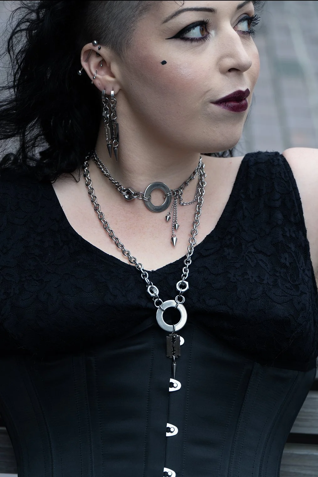 Close-up of a woman with dark makeup and black clothing, wearing multiple silver jewelry chains, hoop earrings, and facial piercings, standing outdoors.