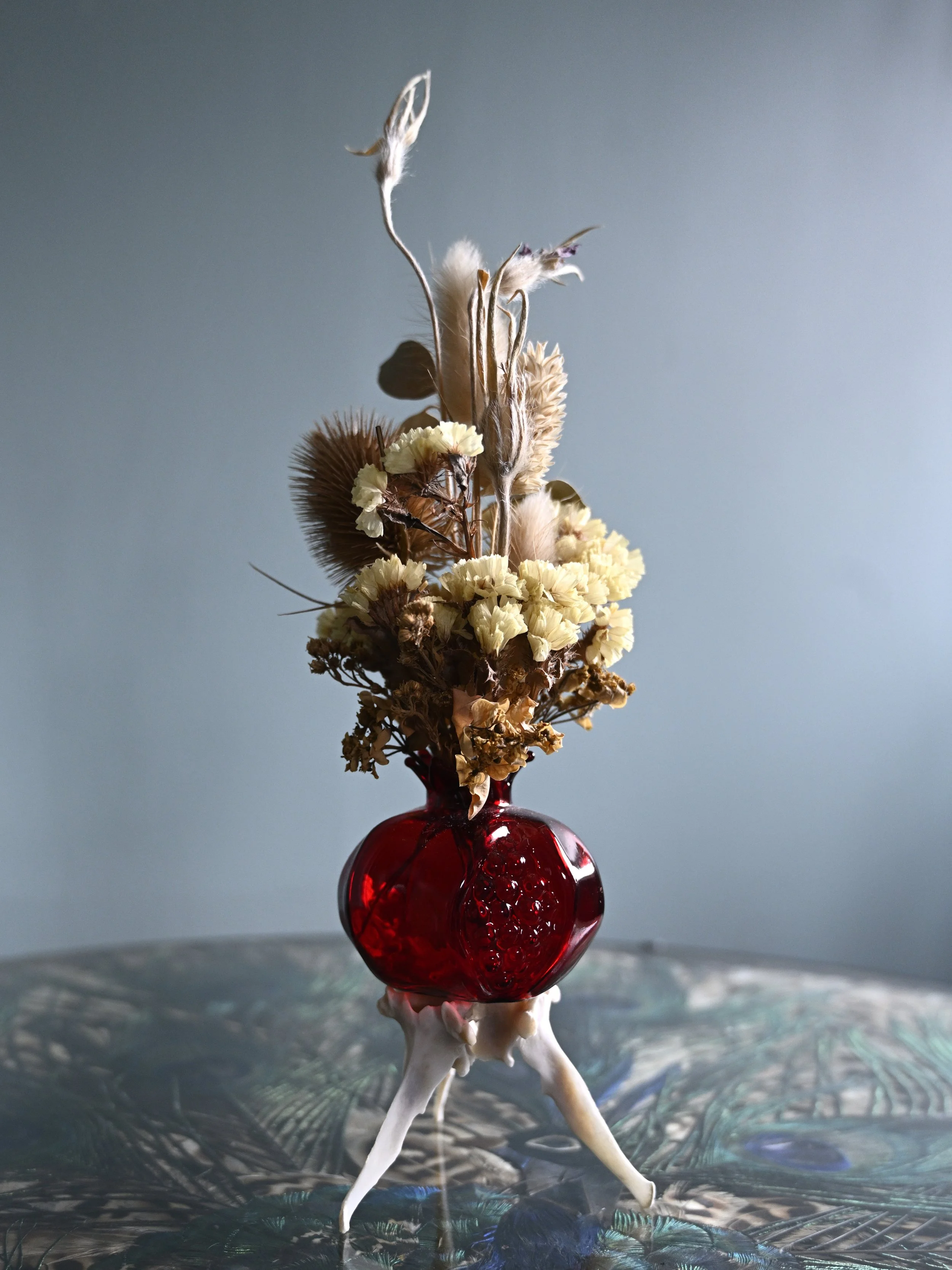 Dried flowers arranged in a red glass vase with uniquely shaped white legs, placed on a reflective glass surface.