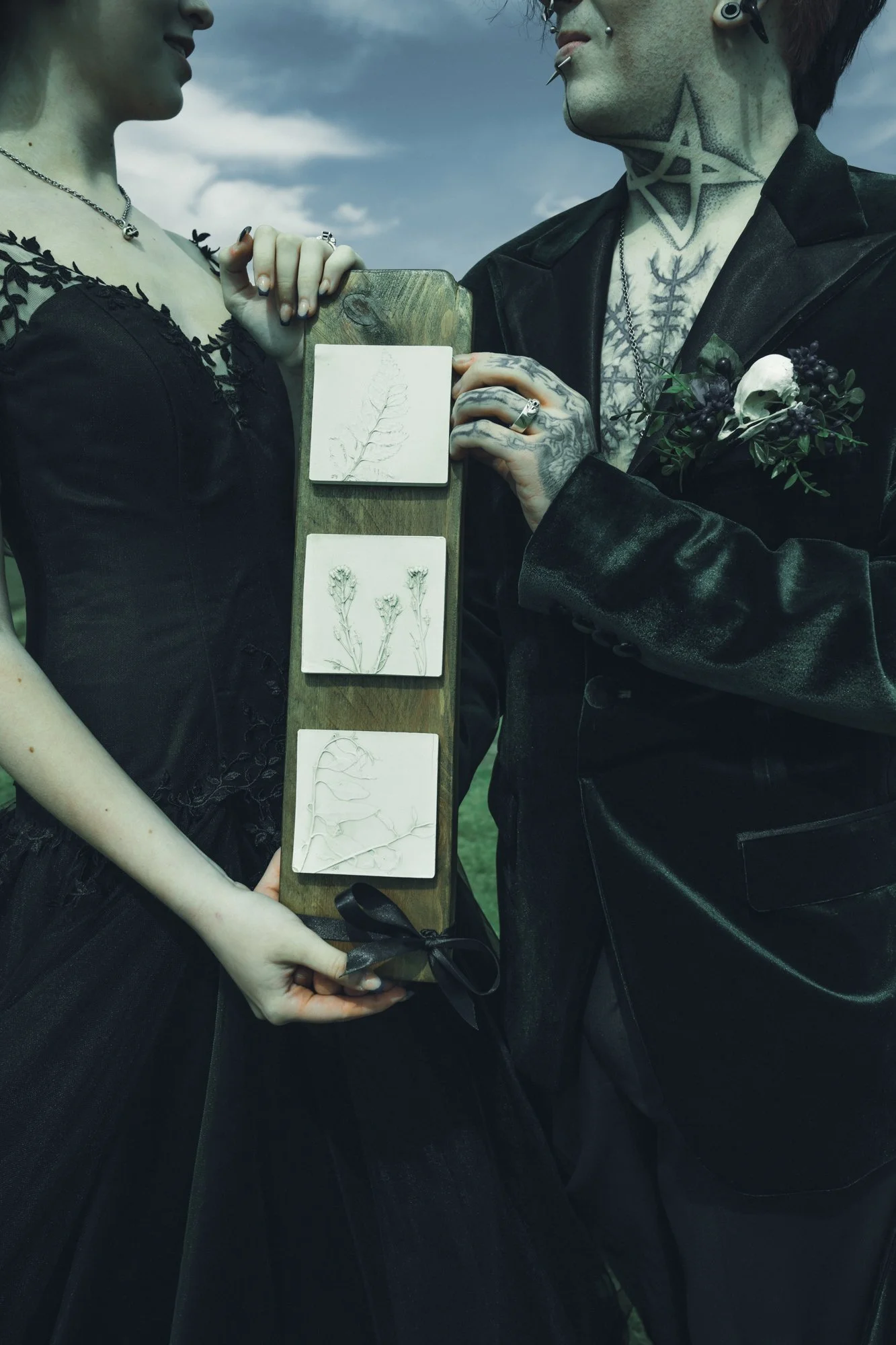 A gothic couple holding a wooden board with three botanical illustrations, standing outdoors against a cloudy sky.