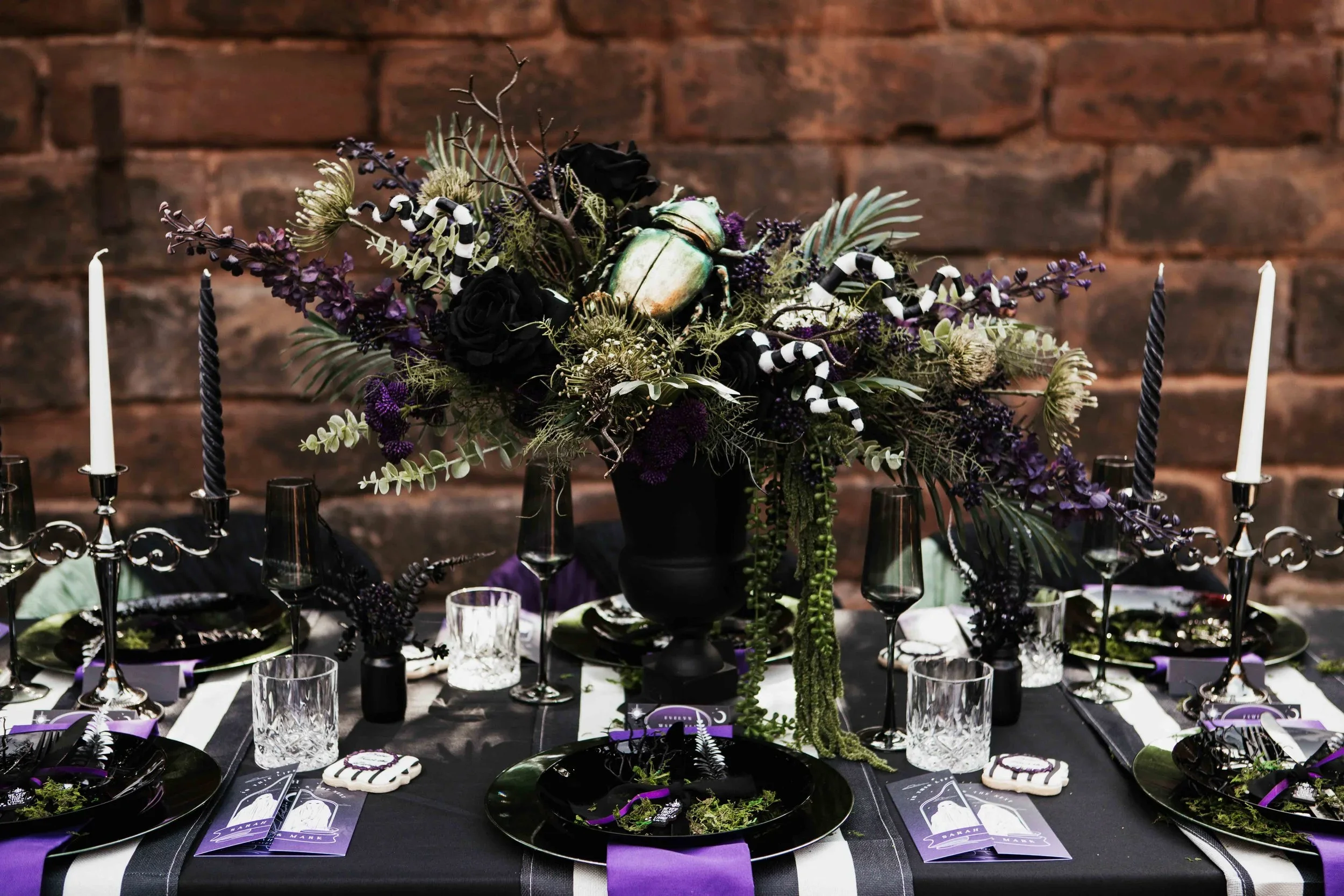 A Halloween themed dining table with black and purple decorations, candles, glassware, and a large floral centerpiece featuring black roses, greenery, and a metallic beetle, against a brick wall background.