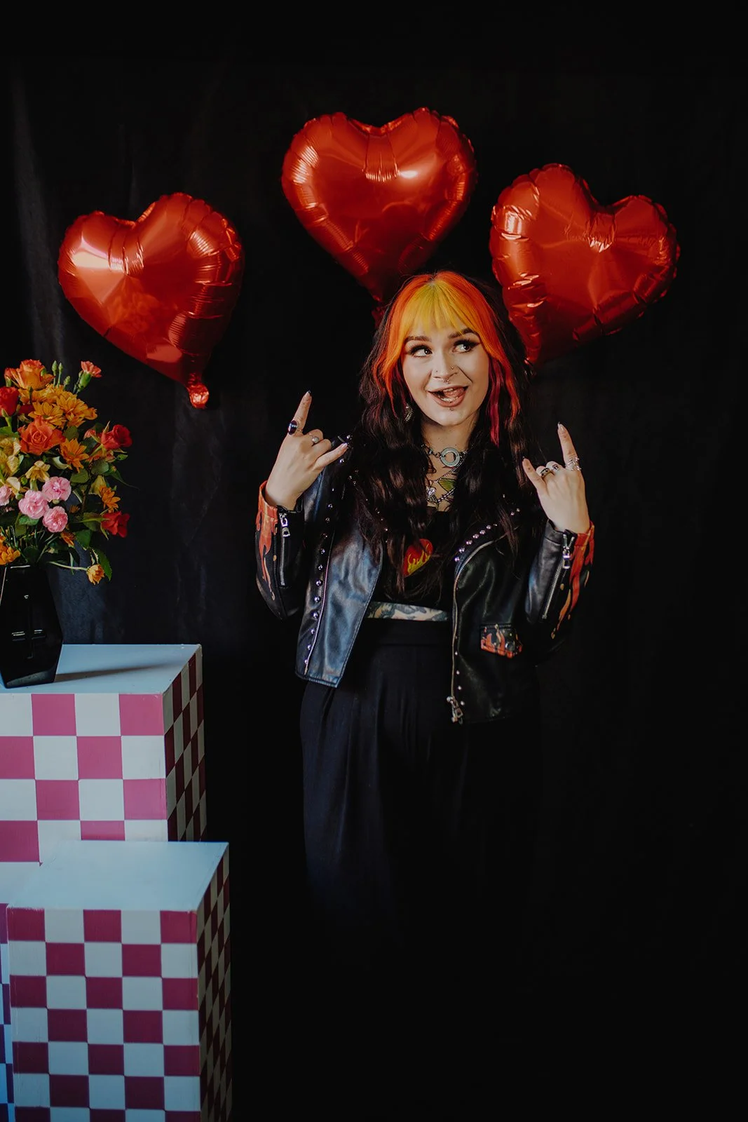 A woman with black and orange hair wearing a leather jacket making rock and roll hand gestures, standing in front of a black background with three red heart-shaped balloons, a bouquet of colorful flowers on a white-and-pink checkered stand.