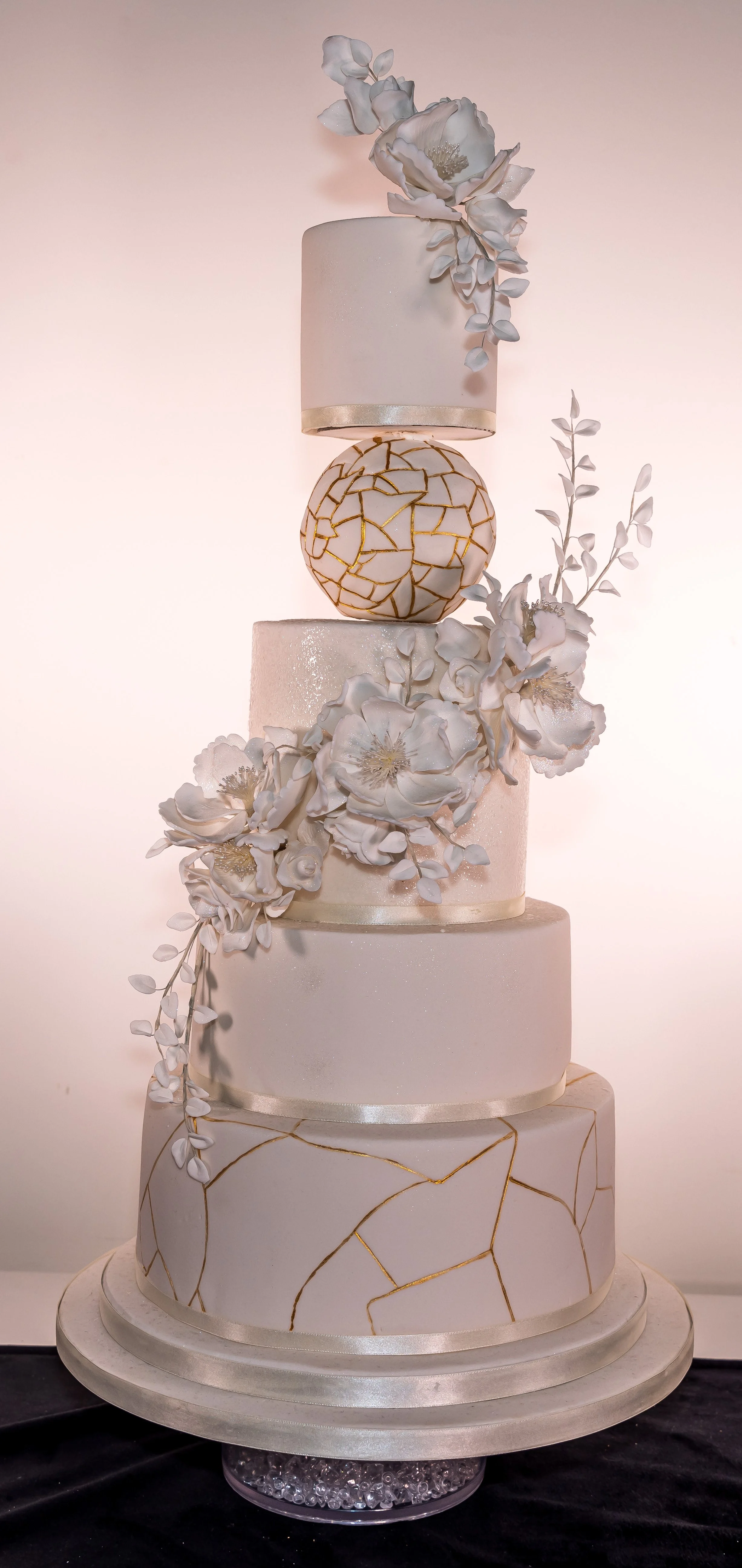 A four-tiered white wedding cake decorated with white flowers and gold accents, displayed on a round cake stand with a beaded base.