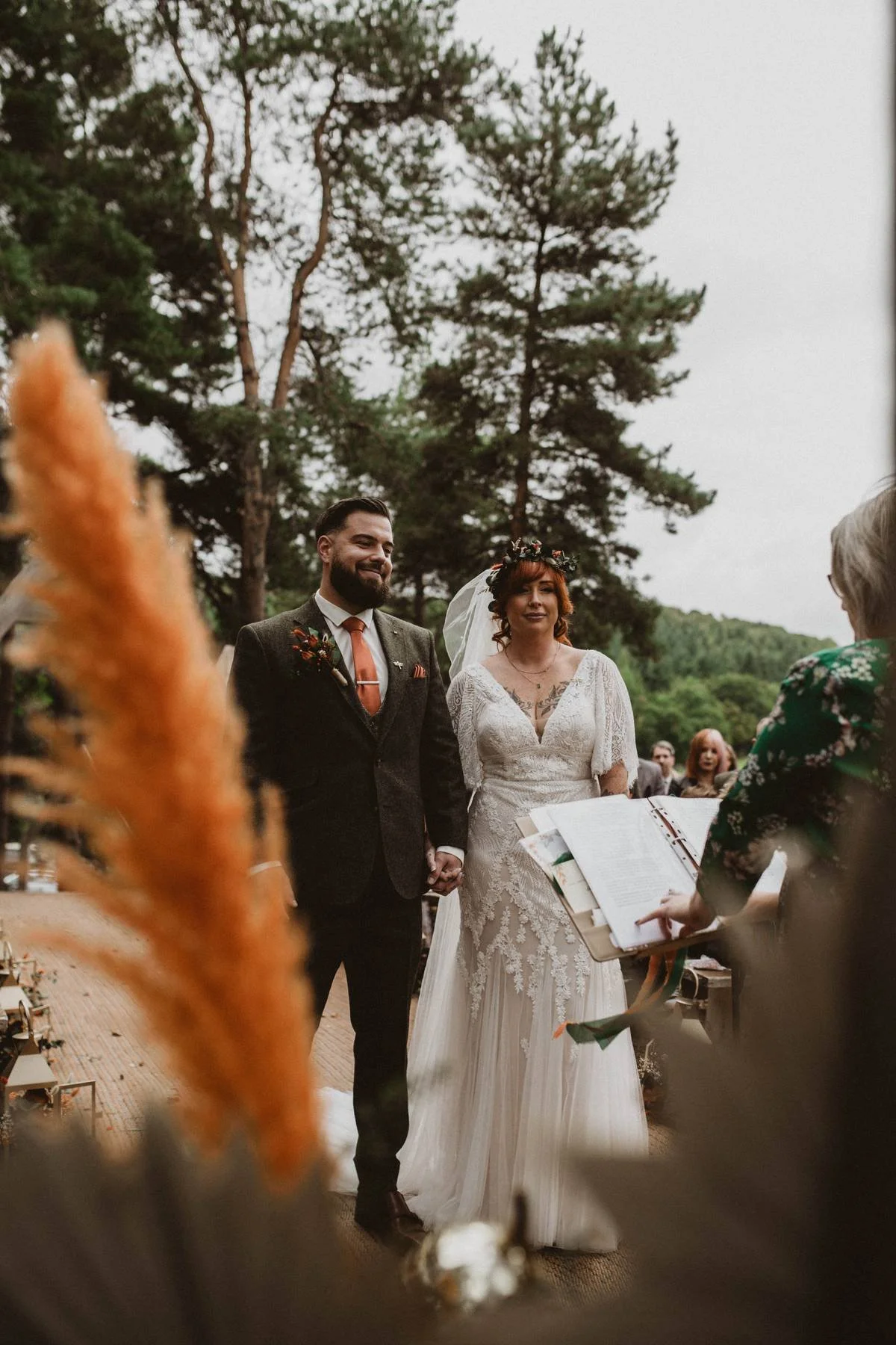 A couple getting married outdoors, holding hands, with a female officiant reading from a book, surrounded by trees and guests, during daytime.