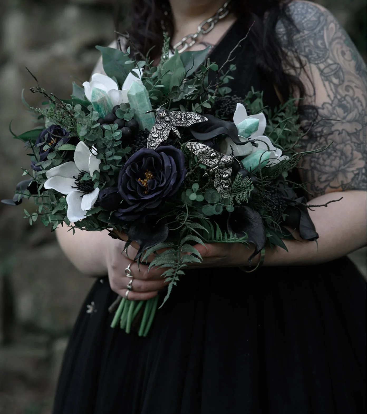 Person holding a bouquet of black, white, and dark purple flowers, with greenery and decorative insects, wearing black gloves, a black dress, and a chain necklace, with a tattooed arm visible.