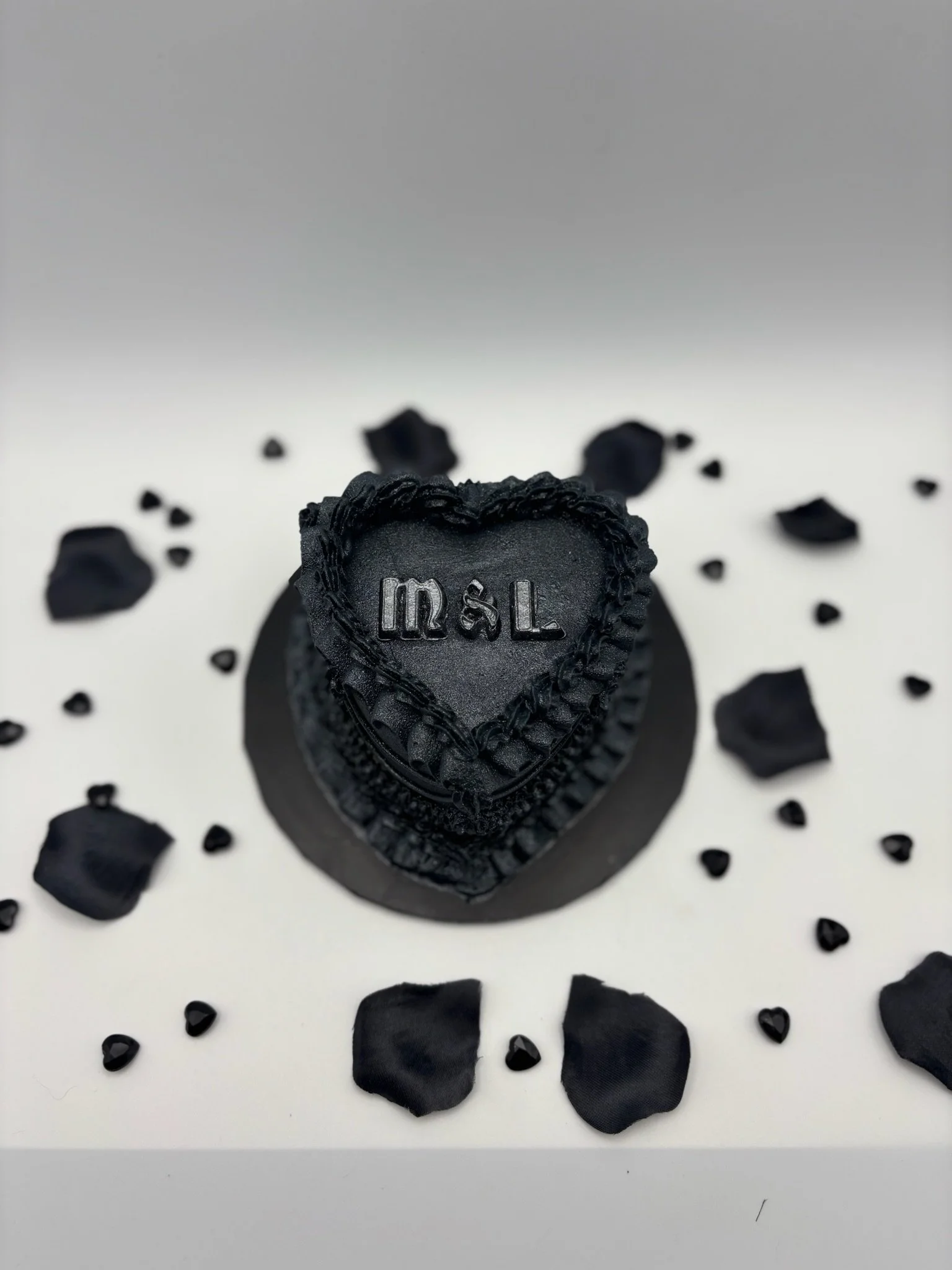A small black heart-shaped cake with decorative piping and the initials 'm&L' on the top, surrounded by black rose petals and small black heart-shaped candies, on a white surface.