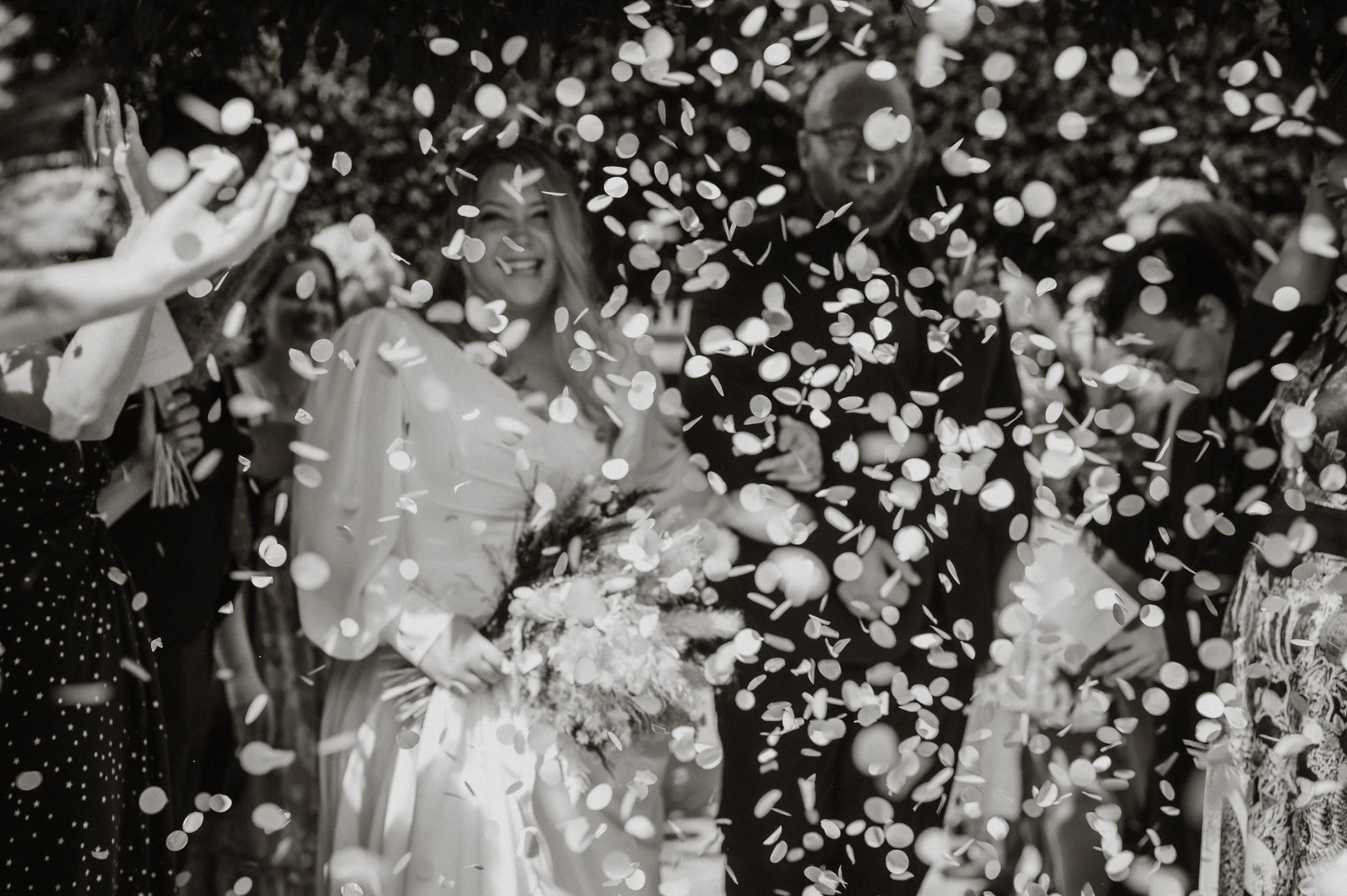 Black and white photo of a wedding celebration with guests throwing confetti around the bride and groom, who are smiling and holding flowers.