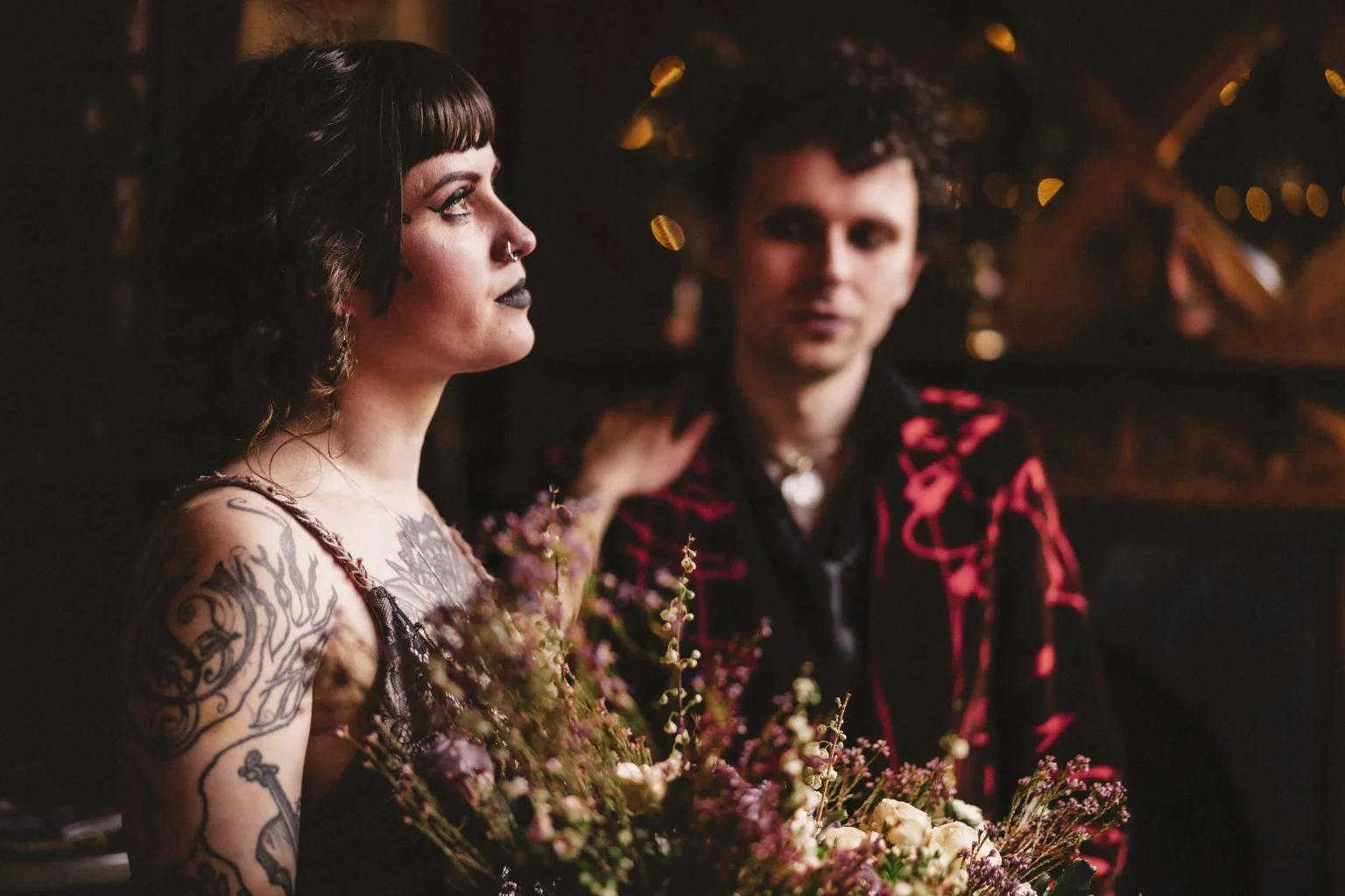 A woman with tattoos on her right arm holding a bouquet of flowers, looking to her left, with a man in a black and red jacket in the background, in a dimly lit setting with warm lights.