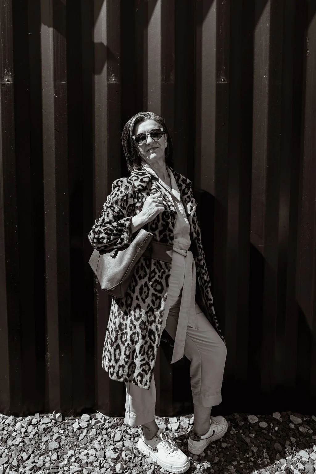 Woman wearing sunglasses and a leopard print coat, holding a handbag, standing outdoors against a dark wooden fence, with gravel on the ground.