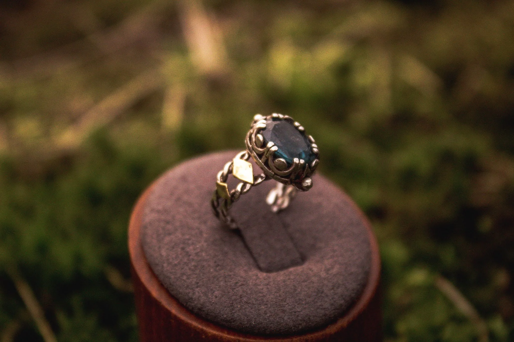 A silver ring with a large dark blue gemstone, set in an ornate silver and gold band, displayed on a cushioned dark gray platform outdoors.