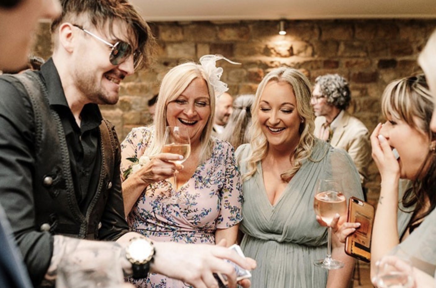 Group of smiling women and a man enjoying a social gathering, holding wine glasses, in a rustic indoor setting with a brick wall background.