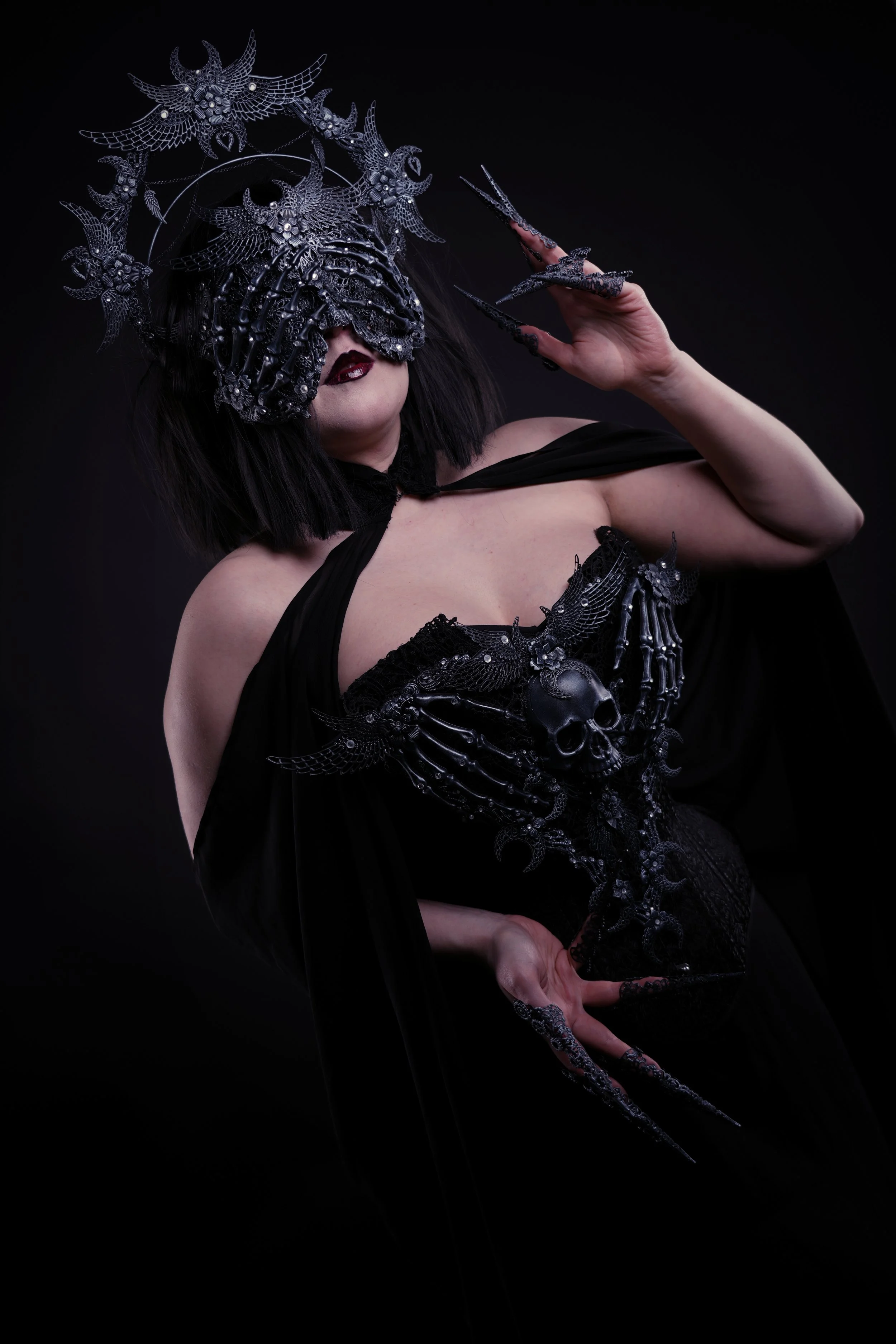 A woman dressed in black with gothic makeup and long dark hair, wearing an elaborate metallic headdress and armor-like clothing adorned with skulls, wings, and skeletal details, against a dark background.