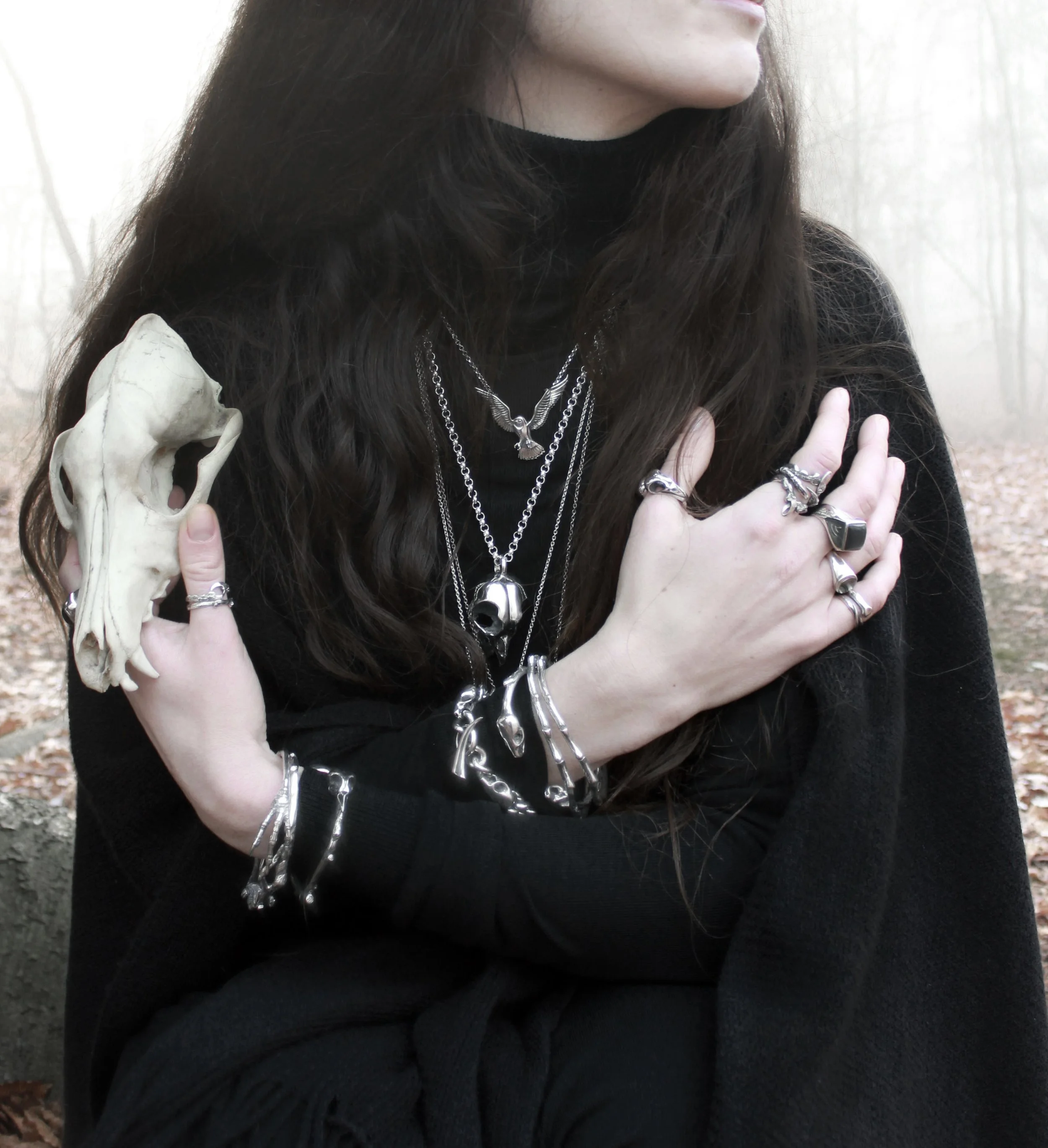 A person with long dark hair wearing multiple silver necklaces and rings, holding a skull, dressed in black, in a foggy forest.