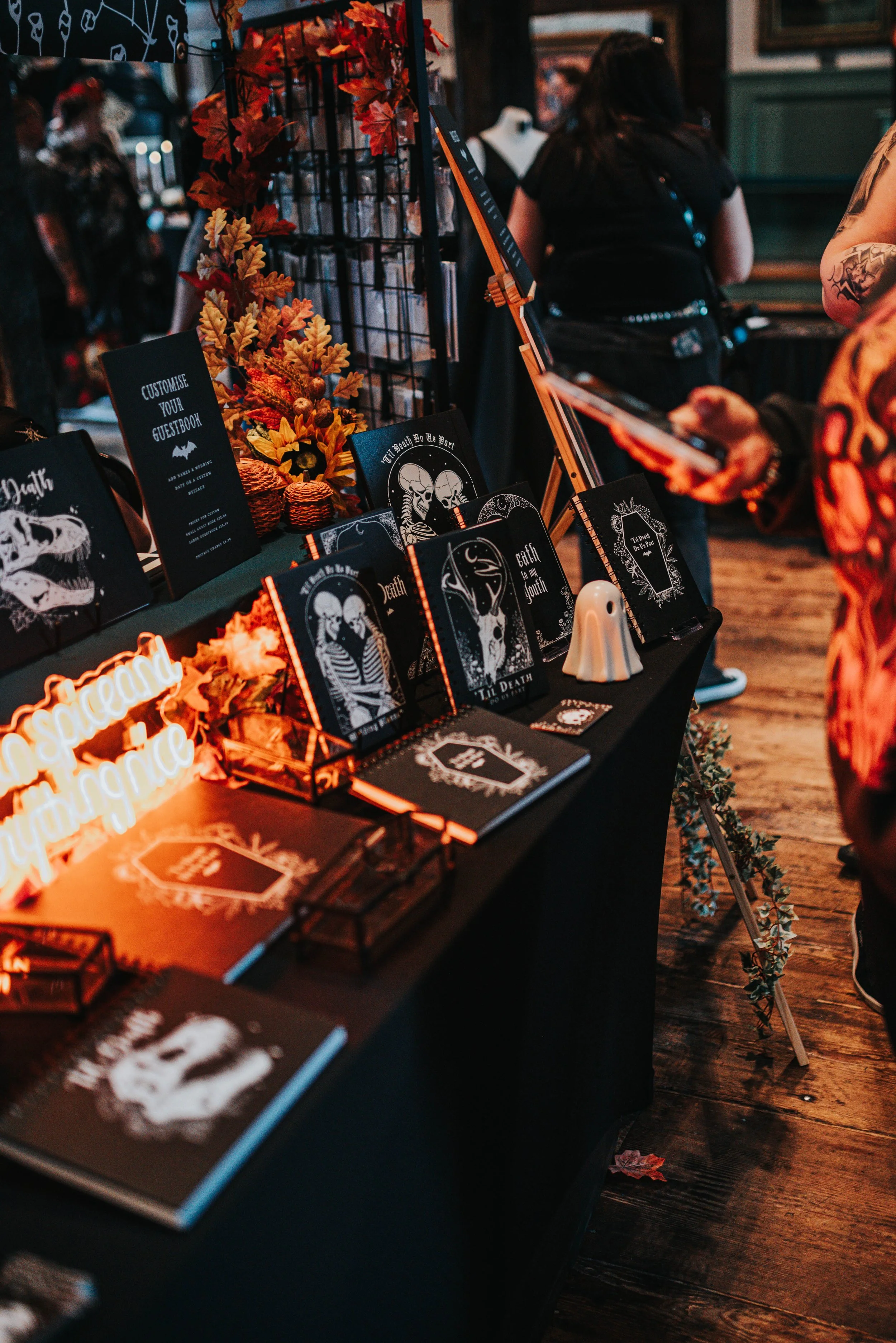A display table at a market or fair decorated with gothic-themed items, including notebooks, signs, and artwork featuring skeletons and skulls, with autumn leaves and glow-in-the-dark signs.